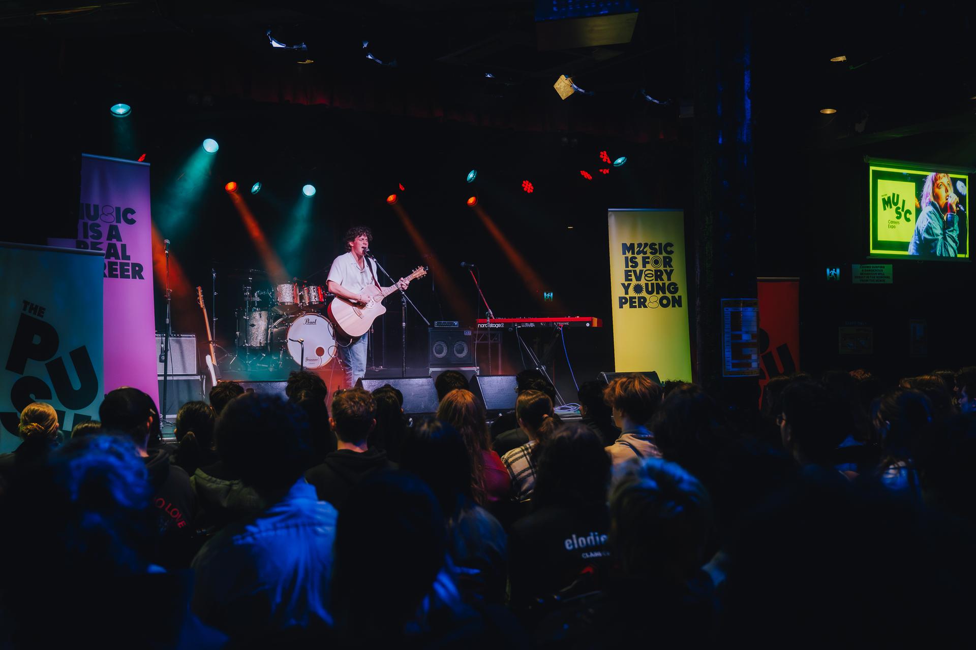 A musician performs with an acoustic guitar on stage under colourful lights, in front of an audience. Banners promoting music are visible.