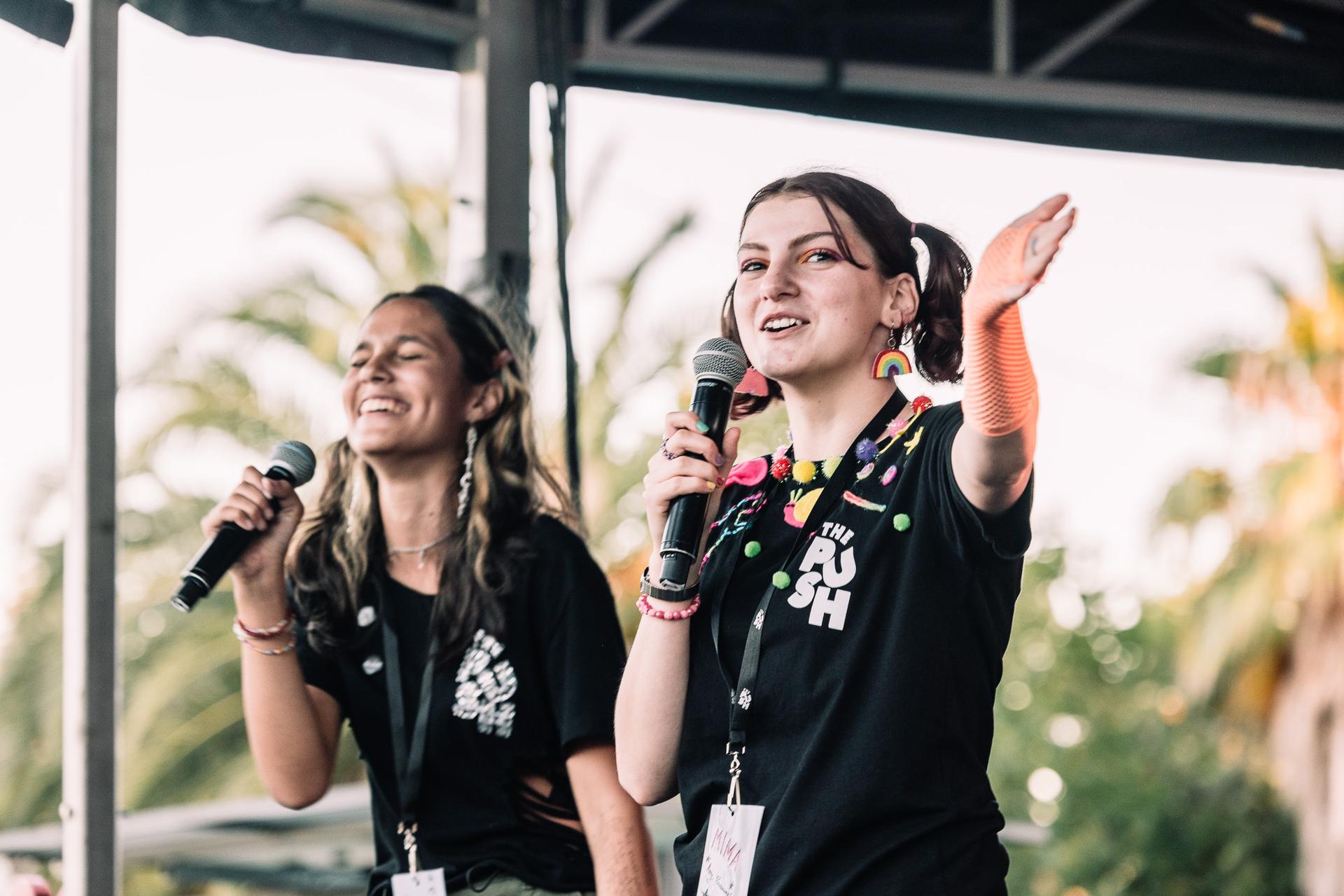 Two young women with microphones performing energetically on an outdoor stage, wearing black shirts and colorful accessories.