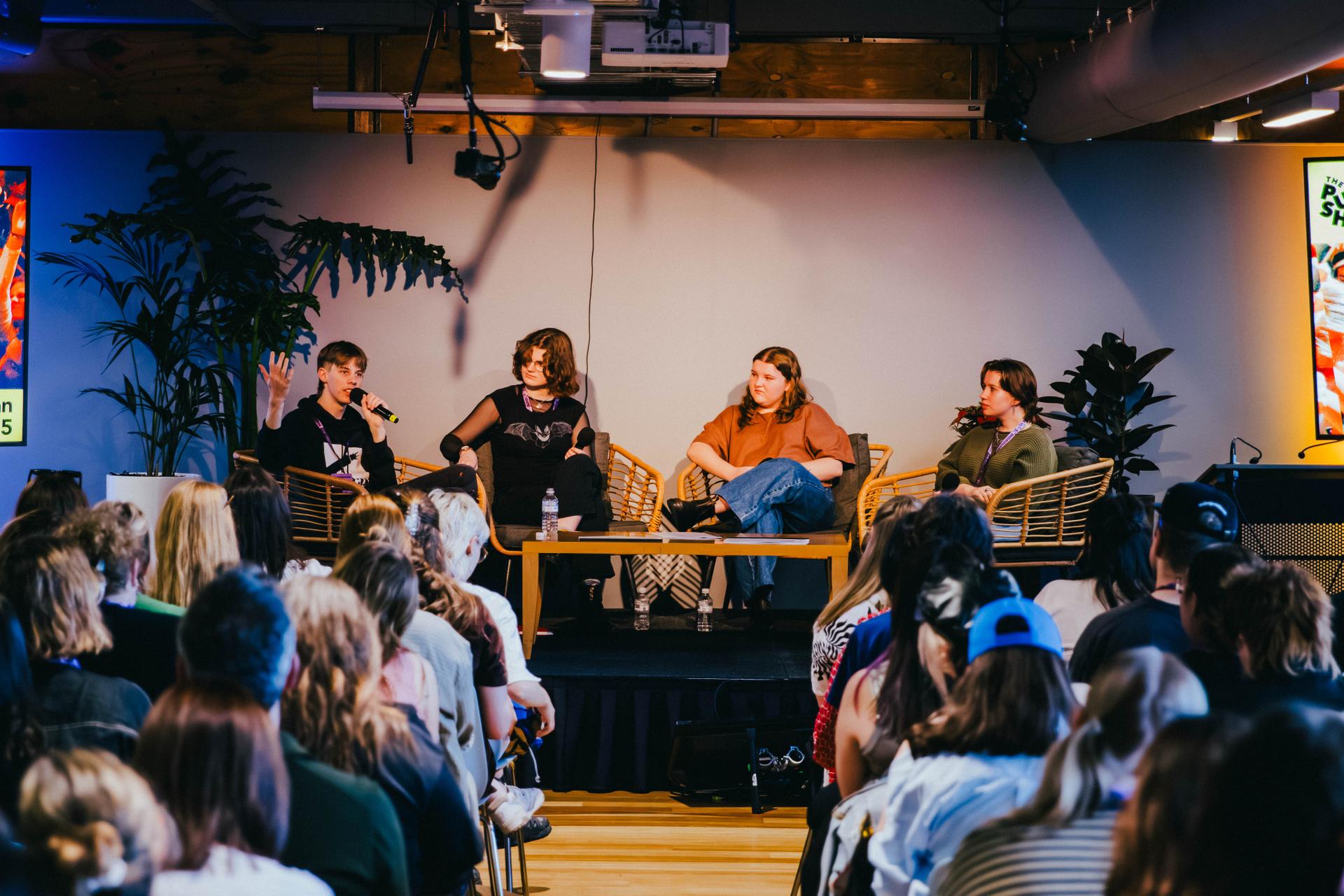 A panel of four people seated on stage, discussing in front of an audience in a modern setting with plants and colorful lighting.