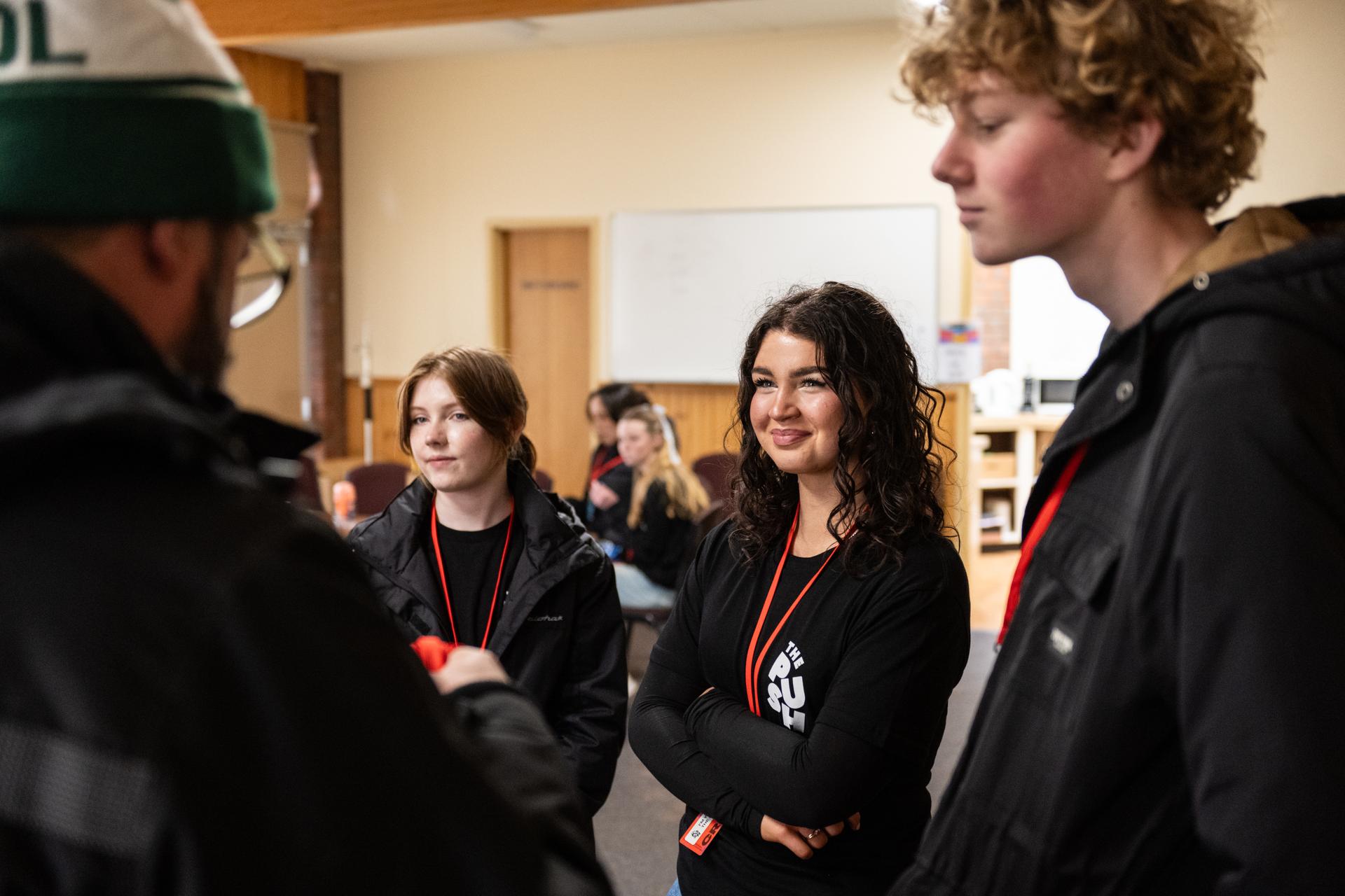A group of young people are standing and listening attentively in a room with a whiteboard in the background.