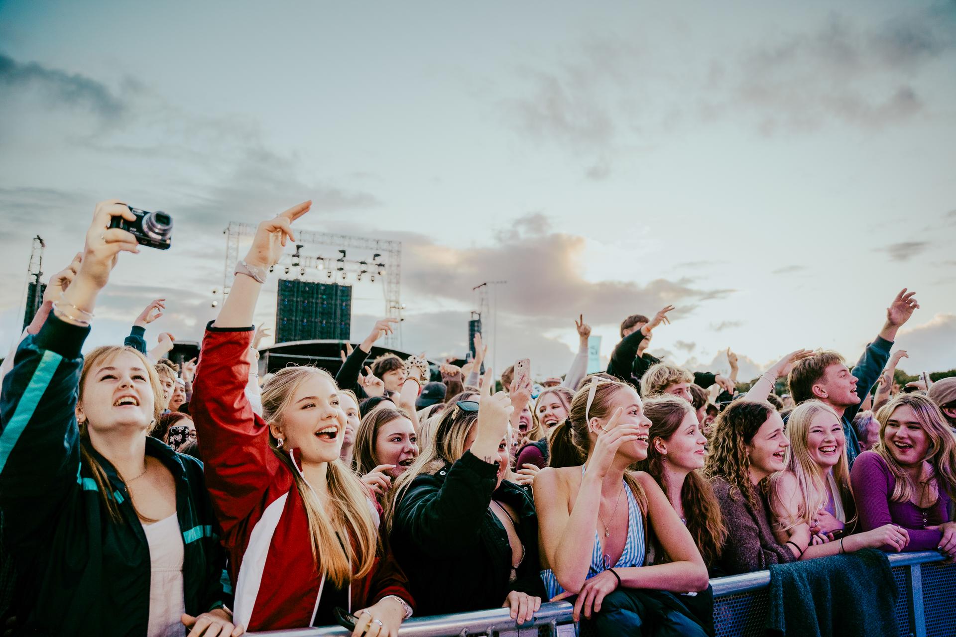 Audience at an outdoor concert, smiling and waving arms, with a backdrop of a cloudy sky.