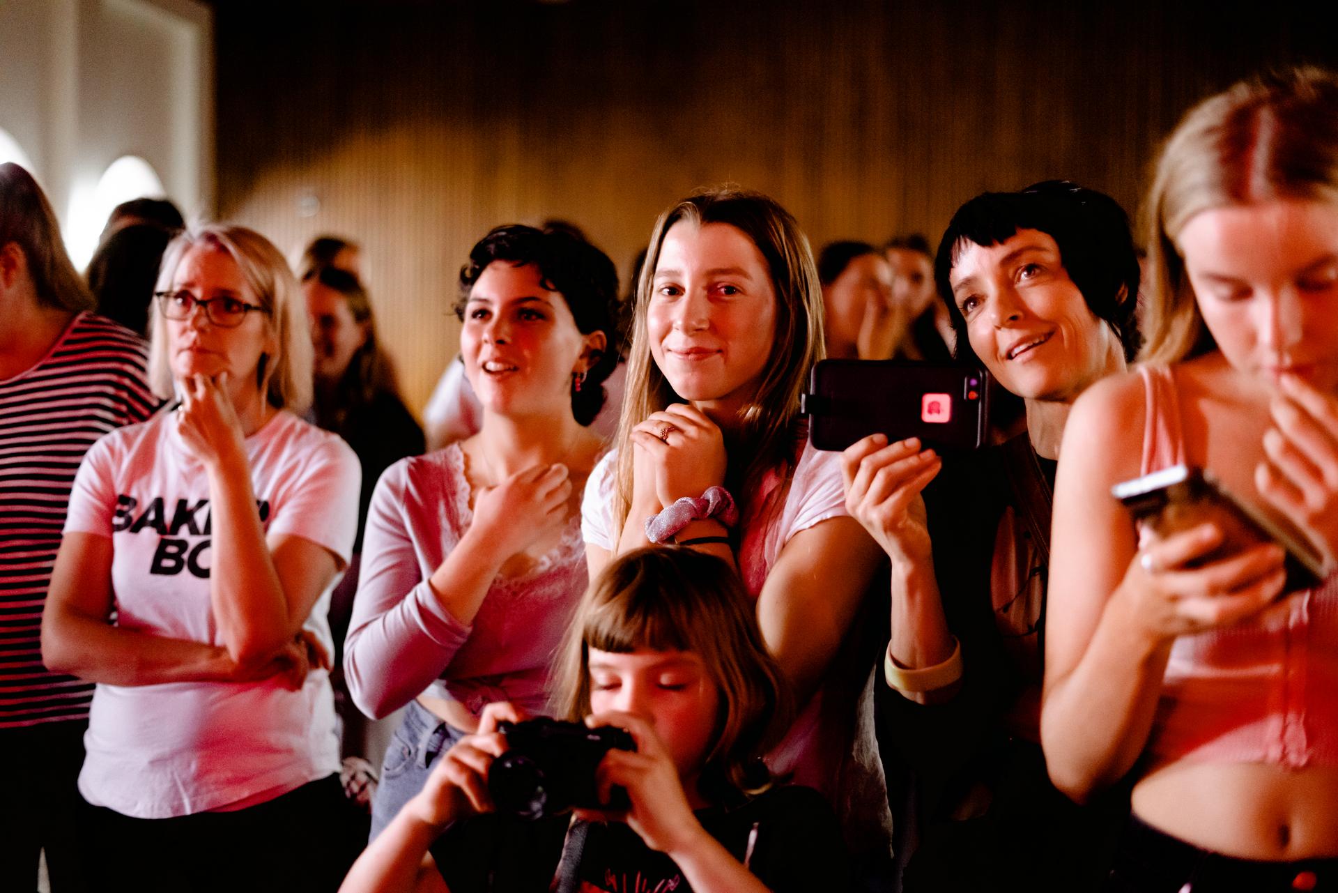 A group of young people stand closely, some holding phones and cameras, eagerly watching something off-camera.