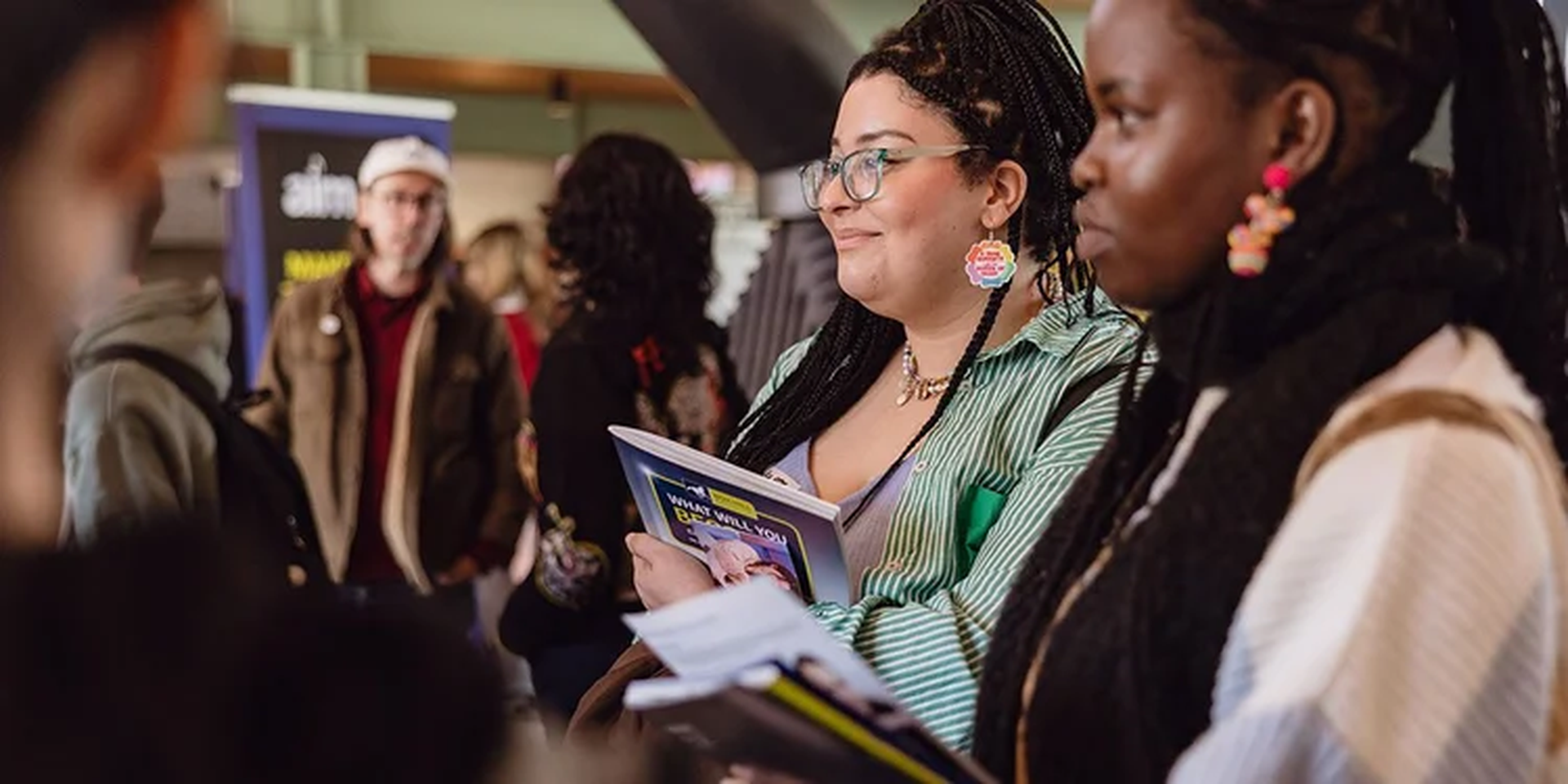 Two people in a lively indoor setting, holding notebooks and pamphlets, with others in the background.