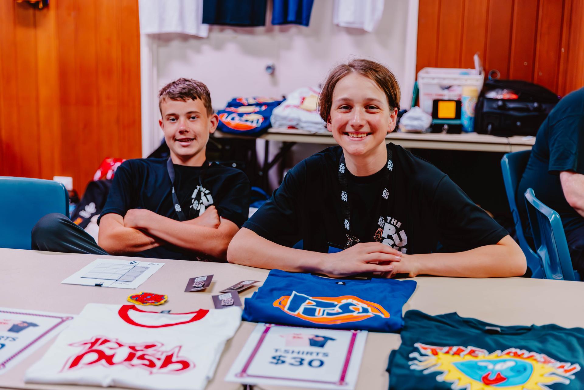 Two smiling individuals sitting behind a table displaying colorful t-shirts and ticket price signs in a casual indoor setting.