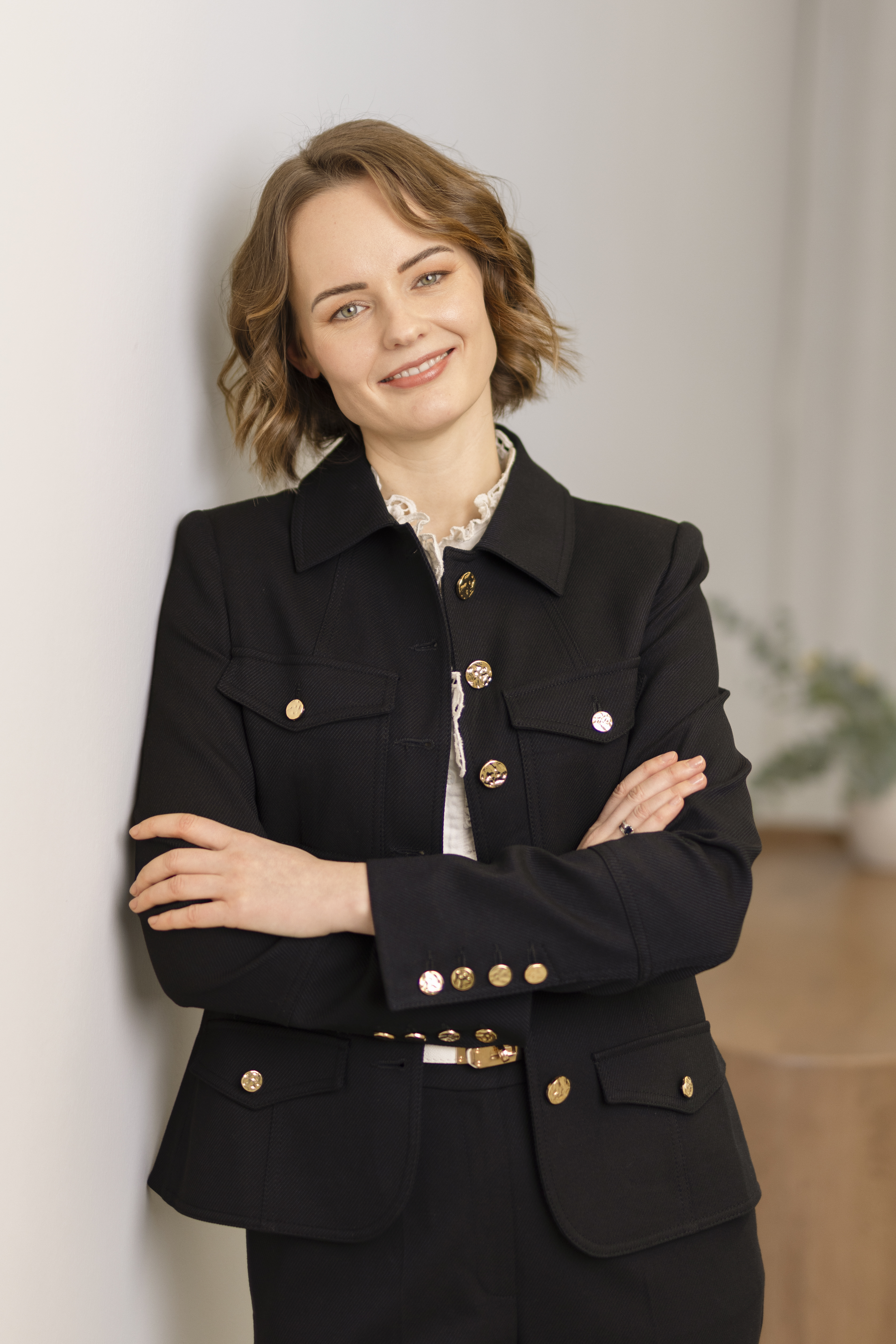 Smiling woman with short wavy hair in a blueblazer, arms crossed, standing against a light-colored wall.