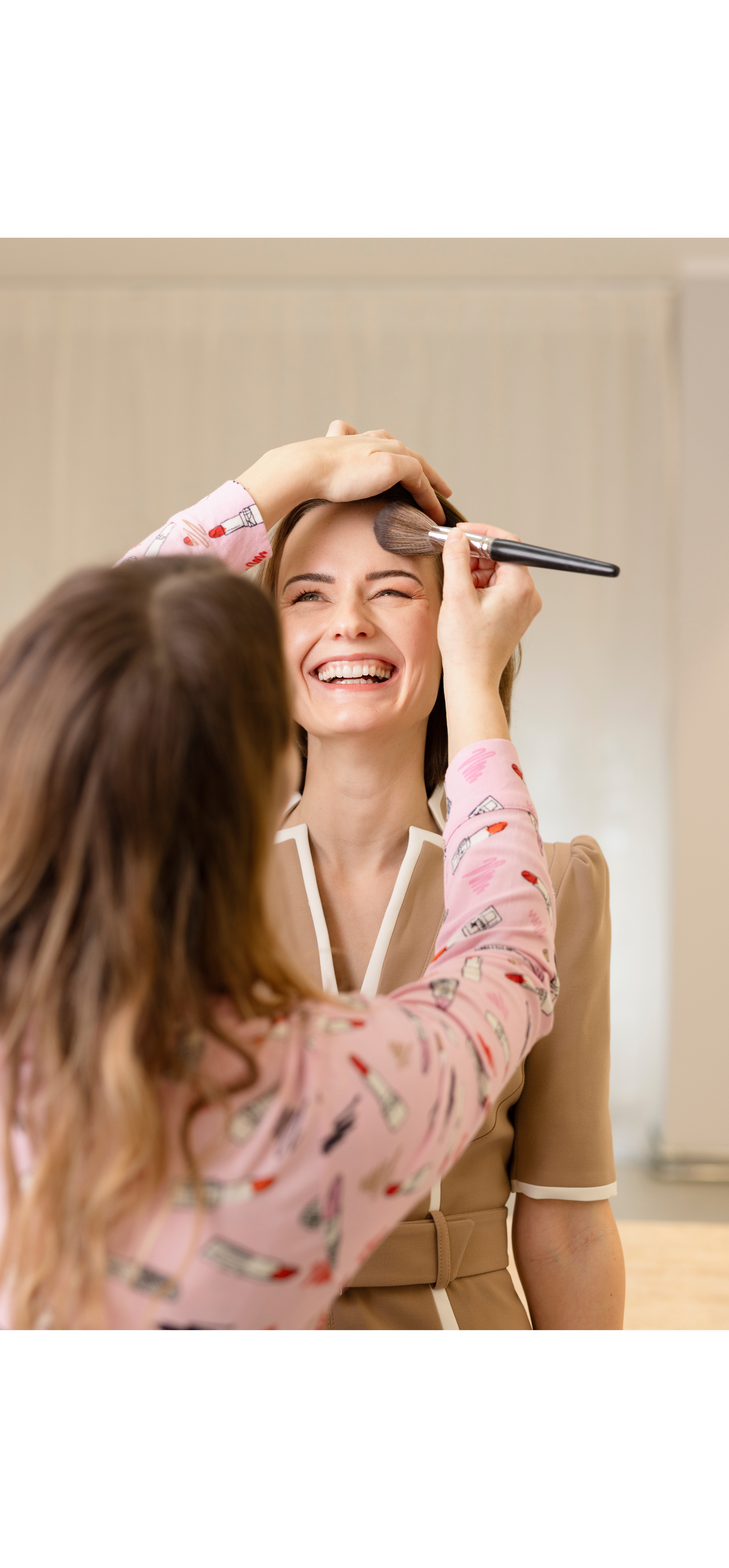 A woman is smiling as another person applies makeup to her forehead with a brush in a bright room.