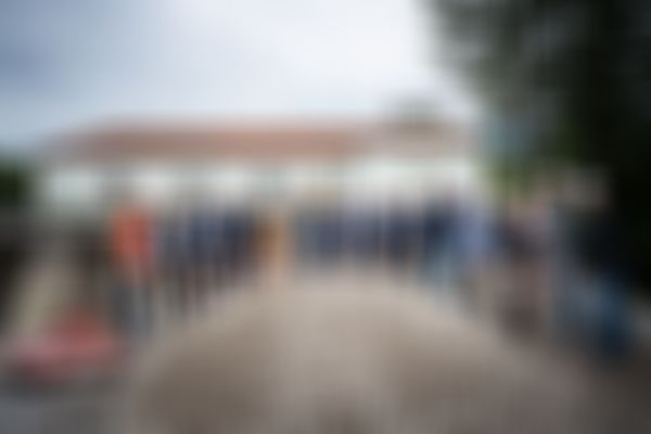 A group of people in hard hats participate in a groundbreaking ceremony, standing with shovels around a mound of gravel.