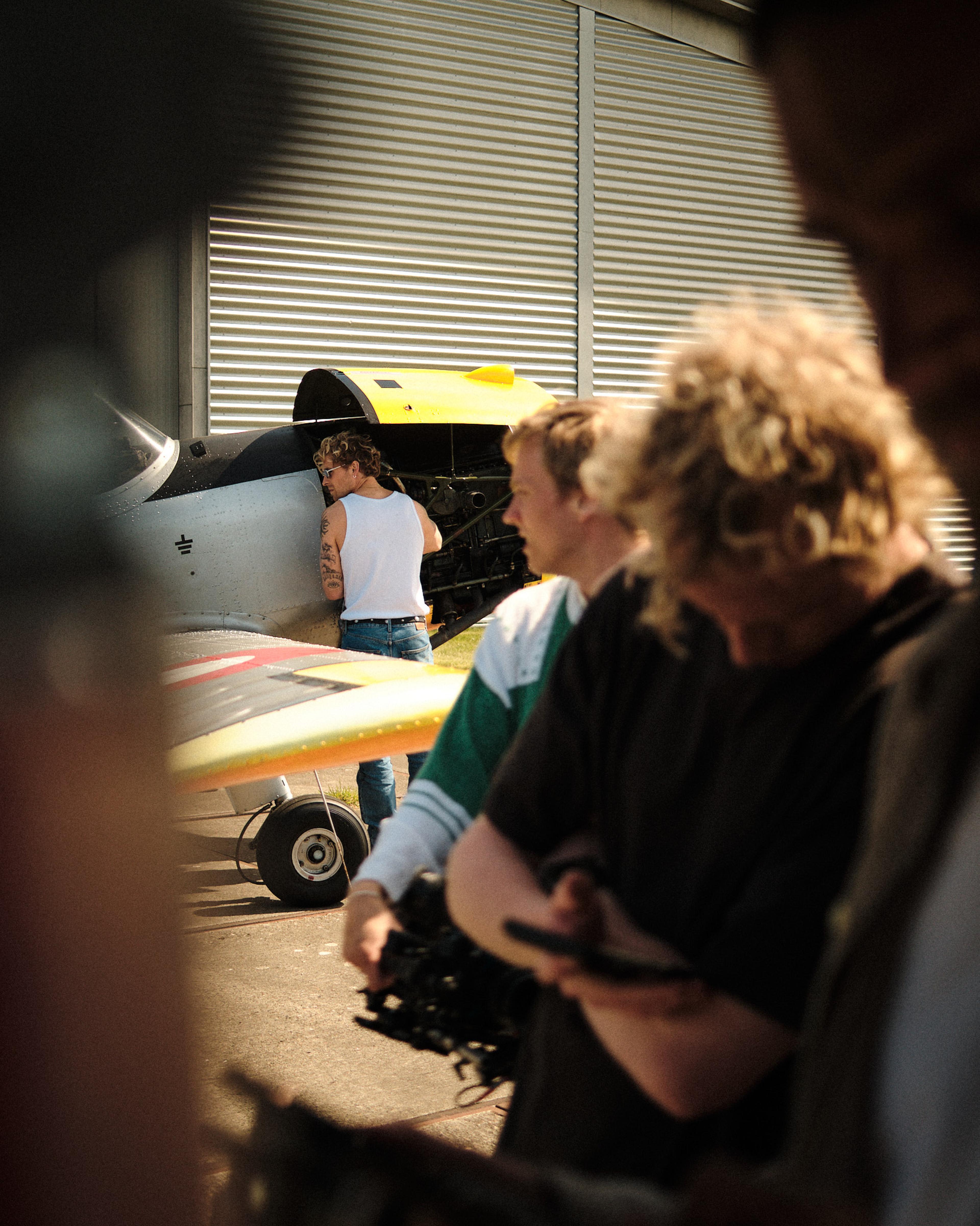 People standing near a small aircraft with an open engine hatch. One man in a tank top and jeans appears to be inspecting it.