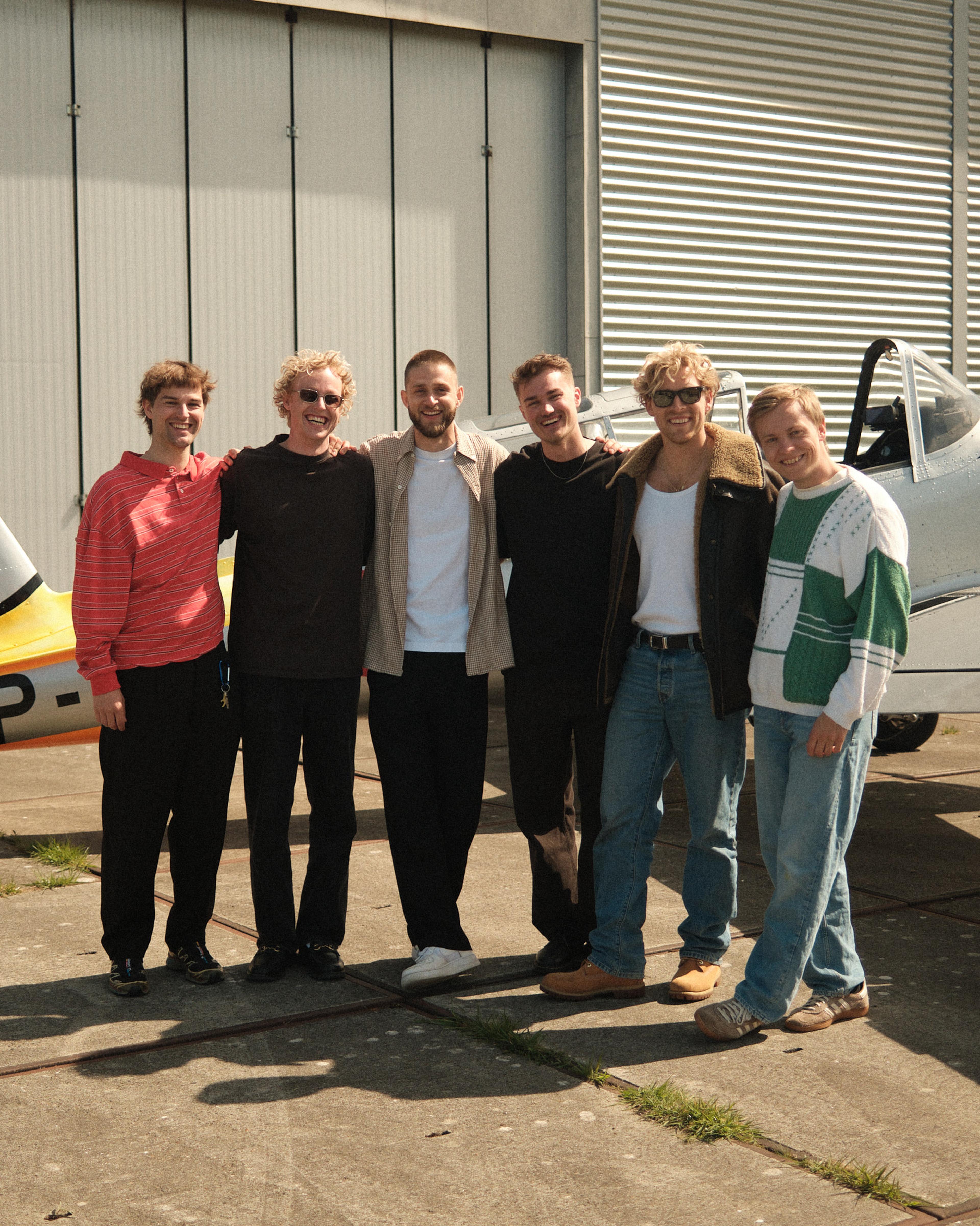 Six men smiling and standing together in front of an airplane hangar on a sunny day.