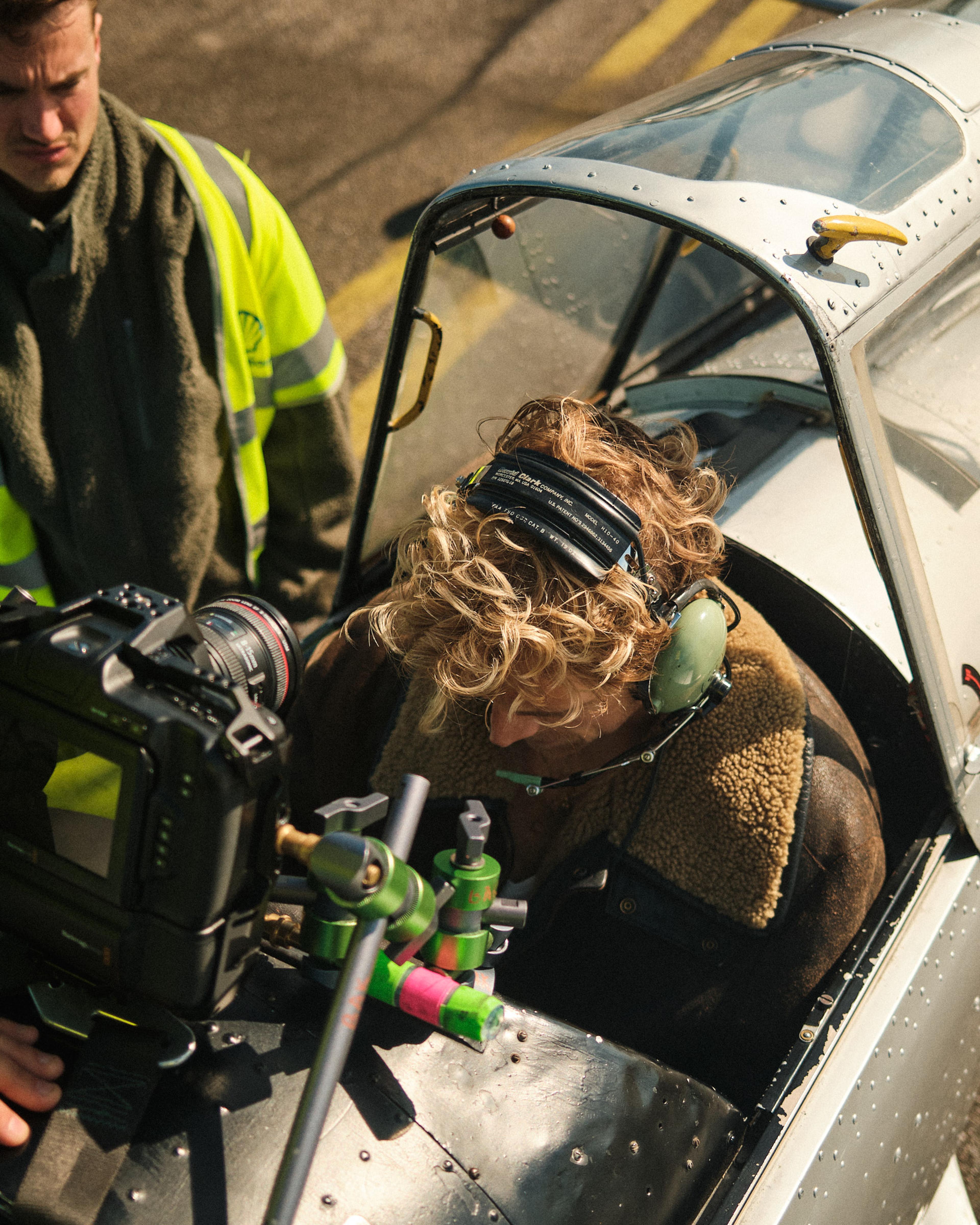 Person in vintage pilot gear sits in an aircraft cockpit, with a camera rig attached. Another person in a high-vis jacket stands nearby.