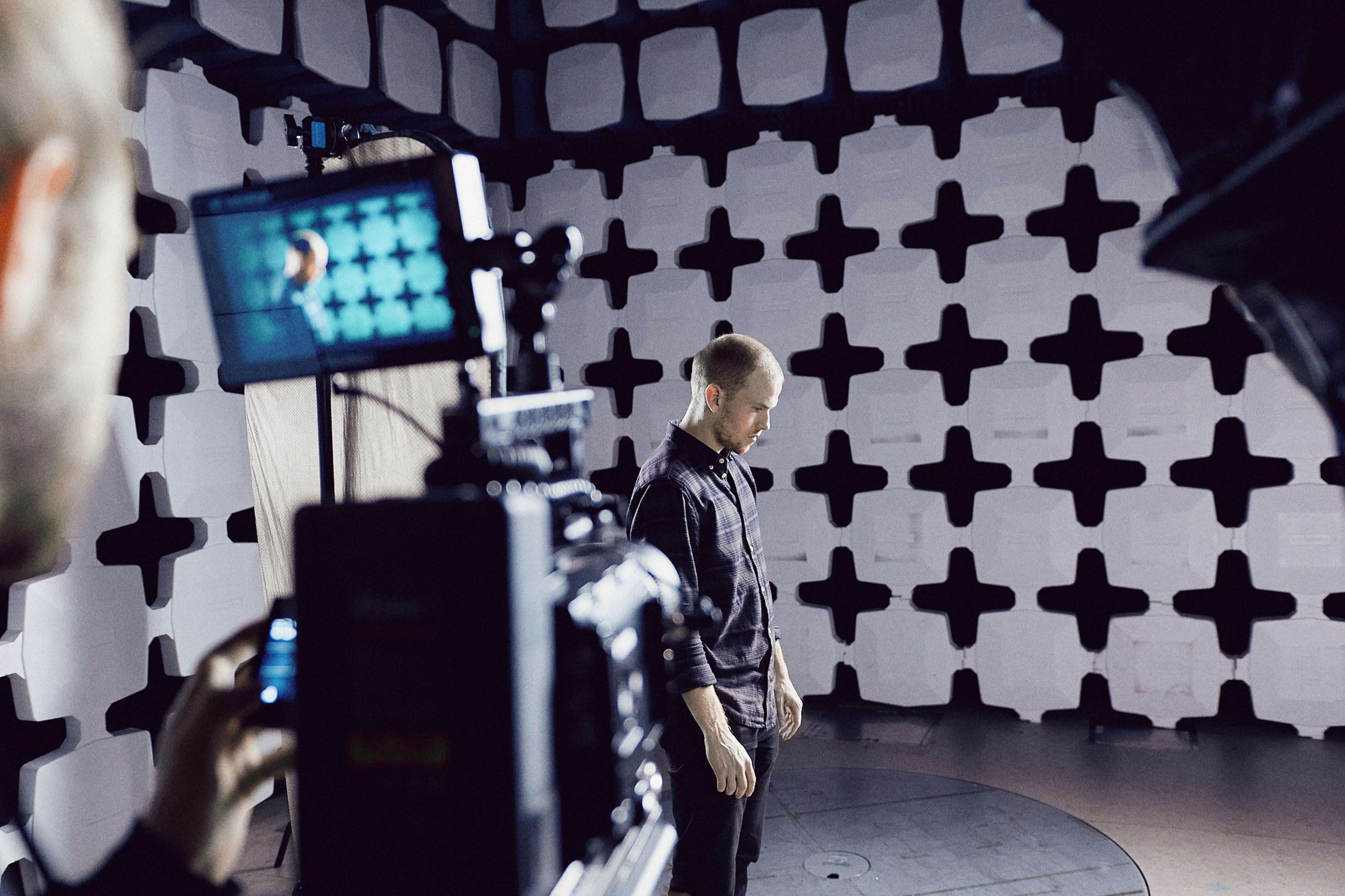 Man stands in anechoic chamber, surrounded by sound-absorbing panels, while being filmed by a camera in the foreground.