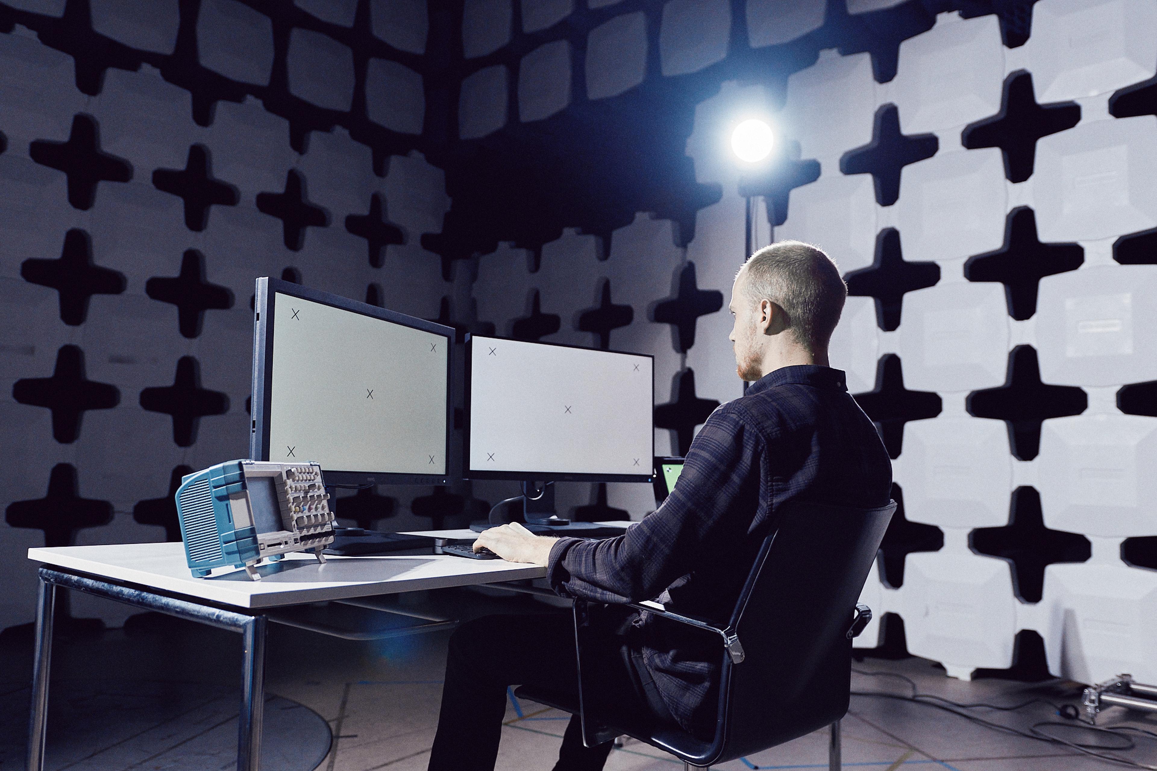 Person working at a desk with three monitors in a room with soundproofing panels and a bright light behind them.