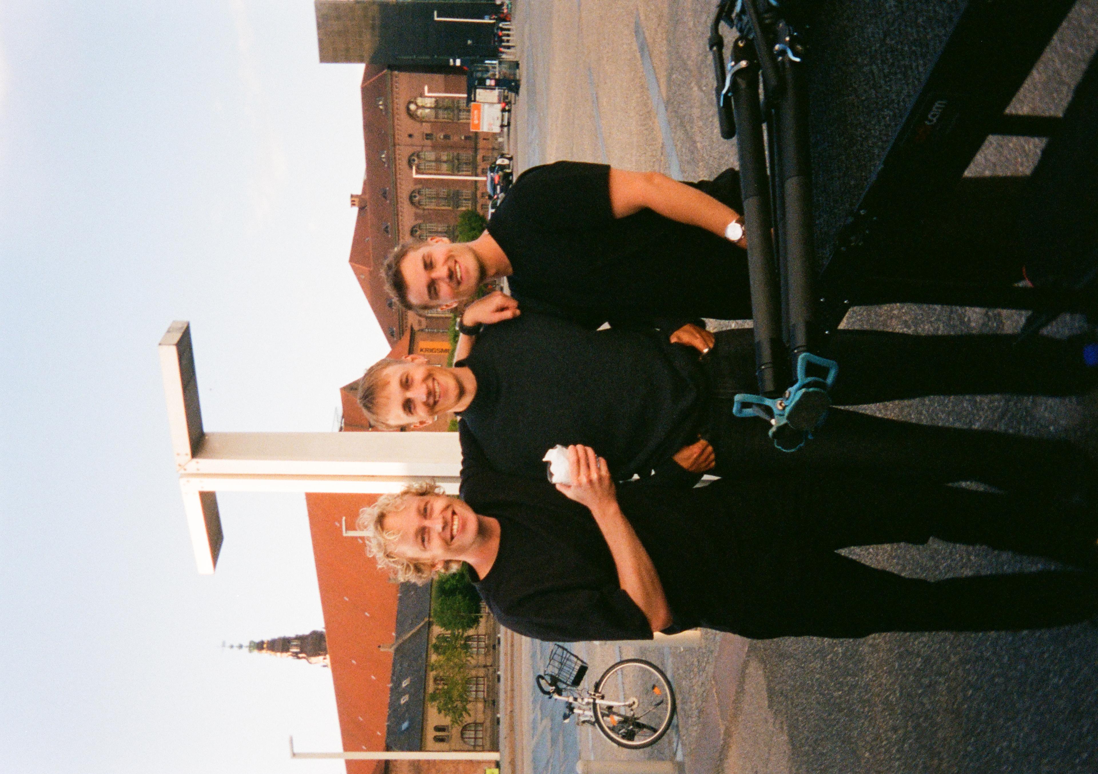 Three smiling men in black outfits stand together outdoors, one holding a drink. Urban background with a bike and historic buildings.