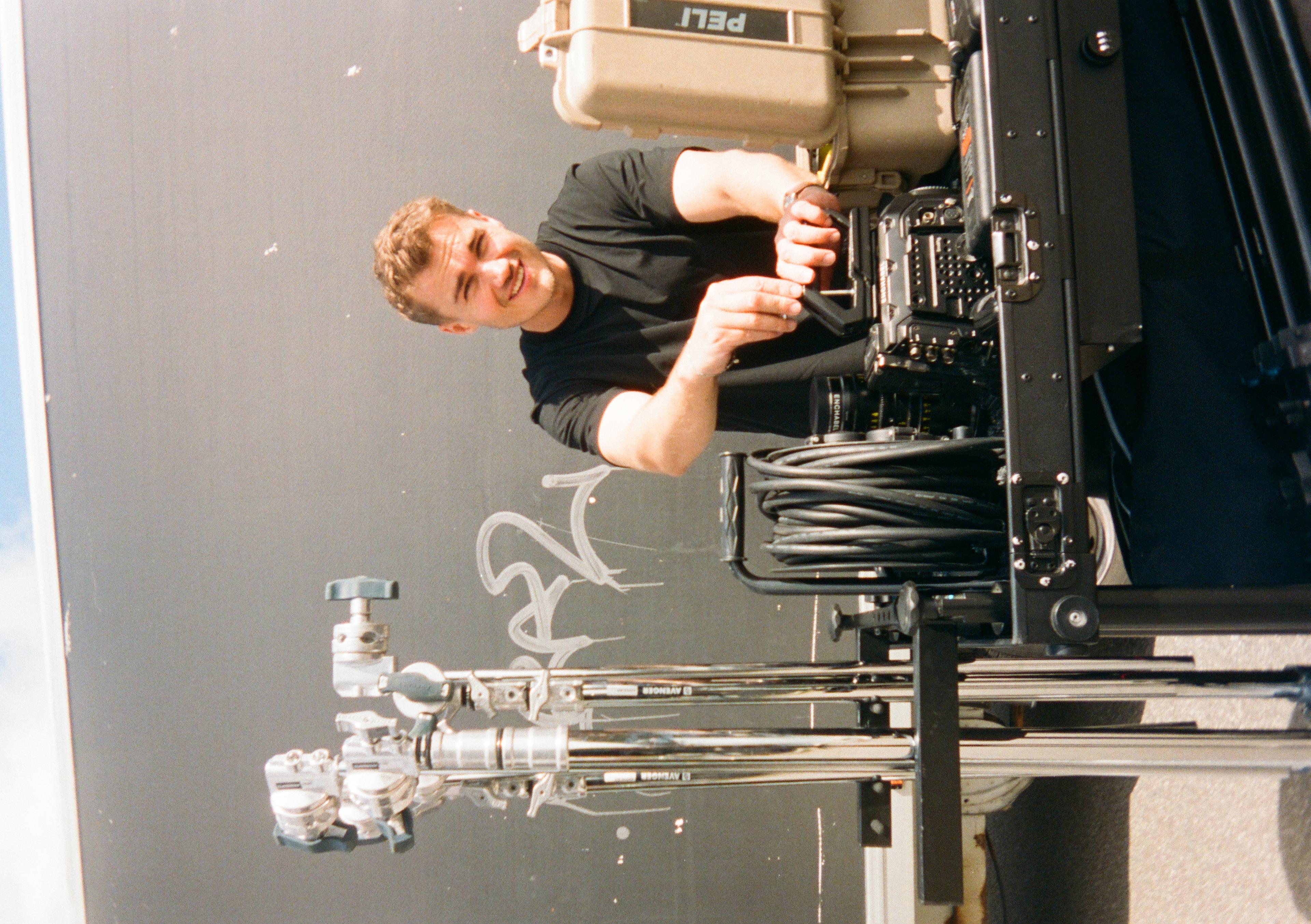 Person smiling, adjusting film equipment, standing next to a truck with gear and cables on a cart. Graffiti on the truck in the background.