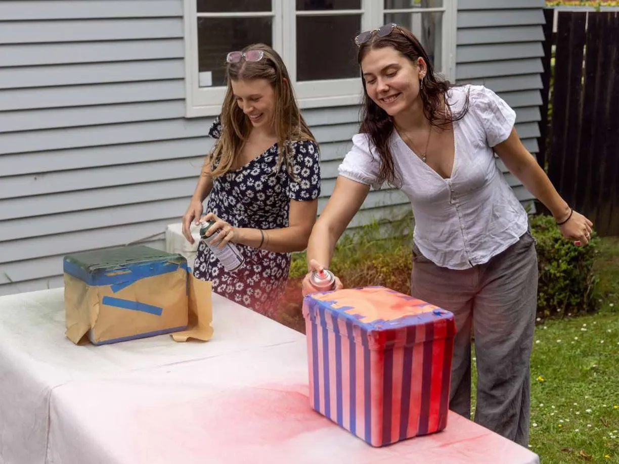 Two women creatively spray-paint striped patterns on boxes outdoors, one with red and the other with blue, near a gray house.