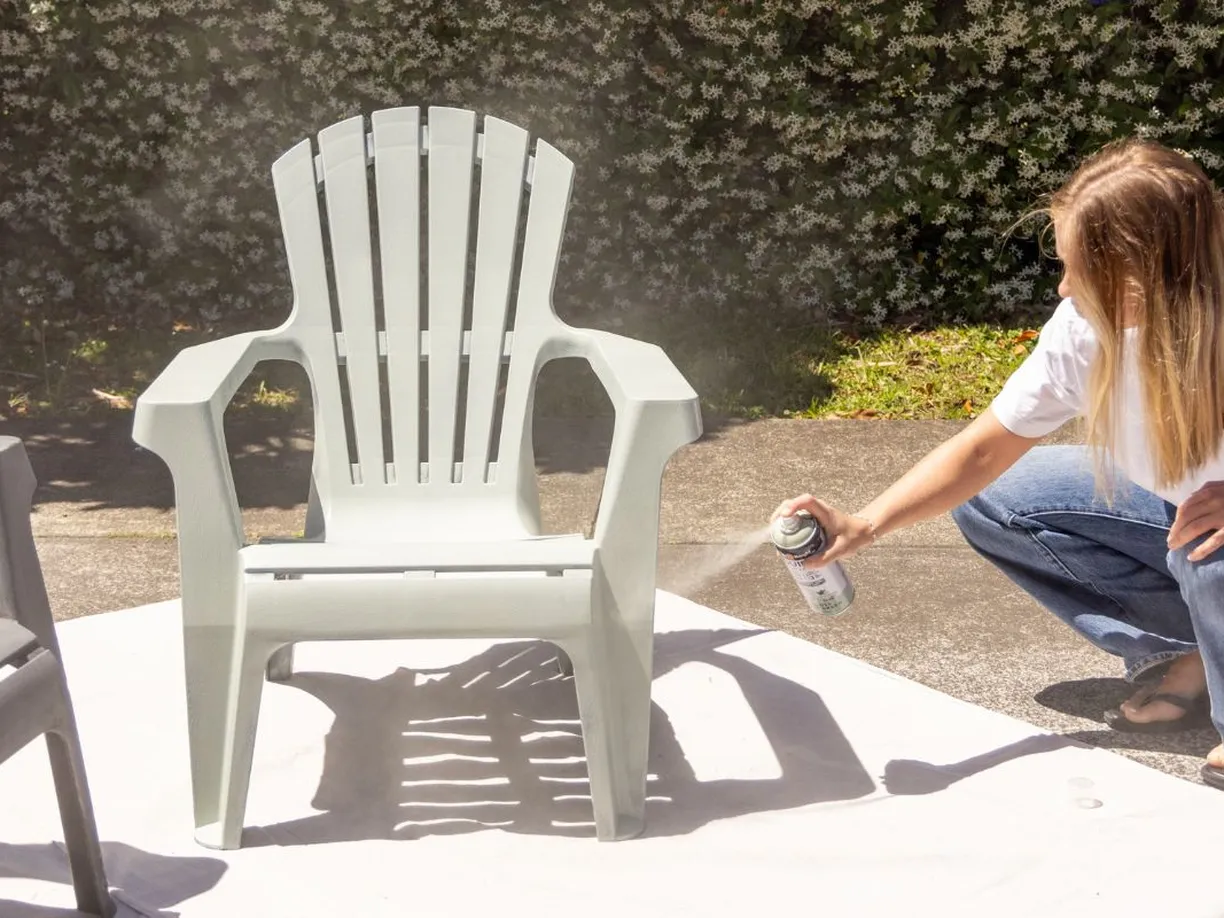 Person spray-painting a white plastic chair outdoors on a sunny day, with greenery in the background.