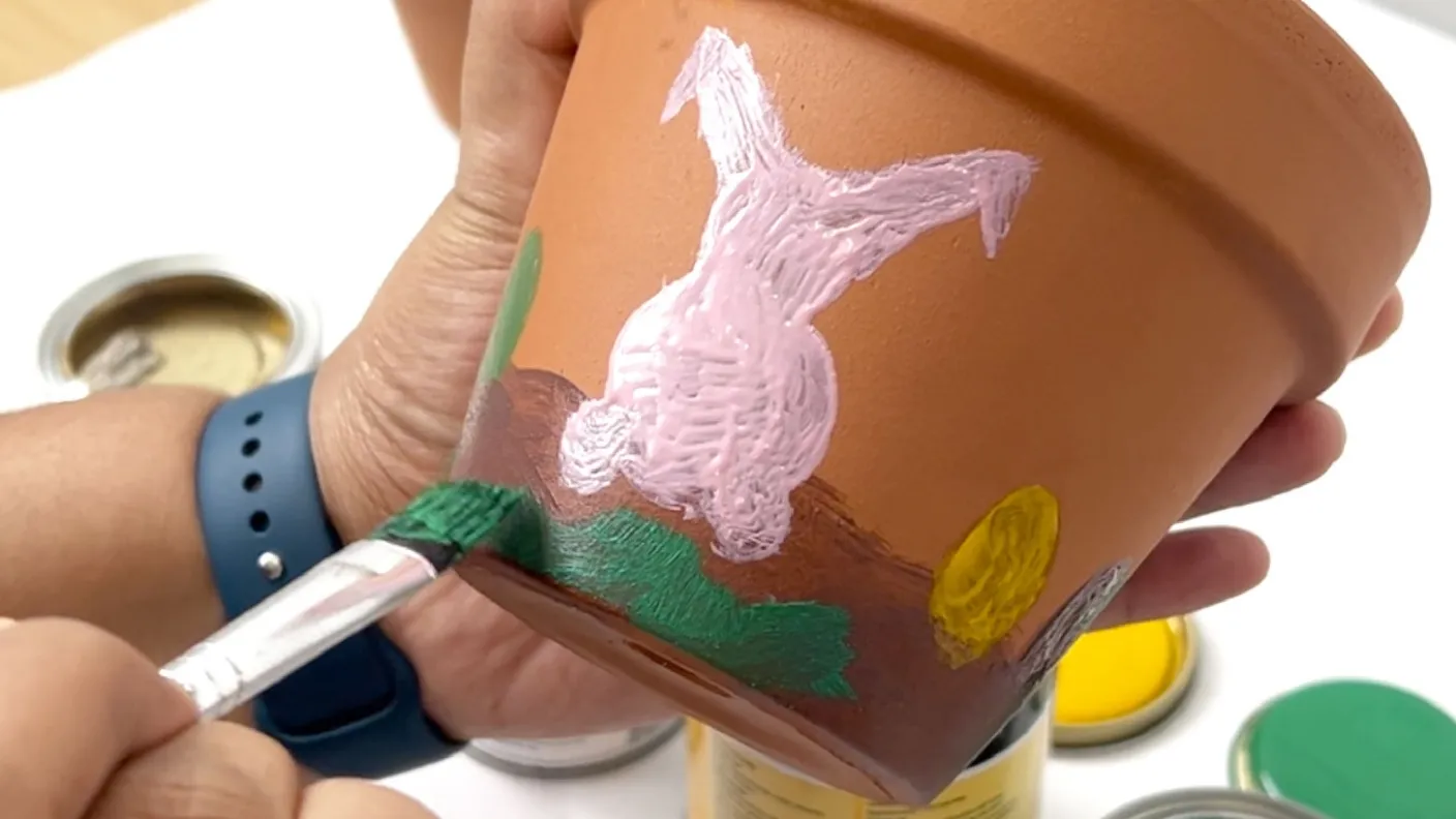 Person painting a terracotta pot with a brush, adding green, pink, and yellow colors. The pot rests on one hand as they work.