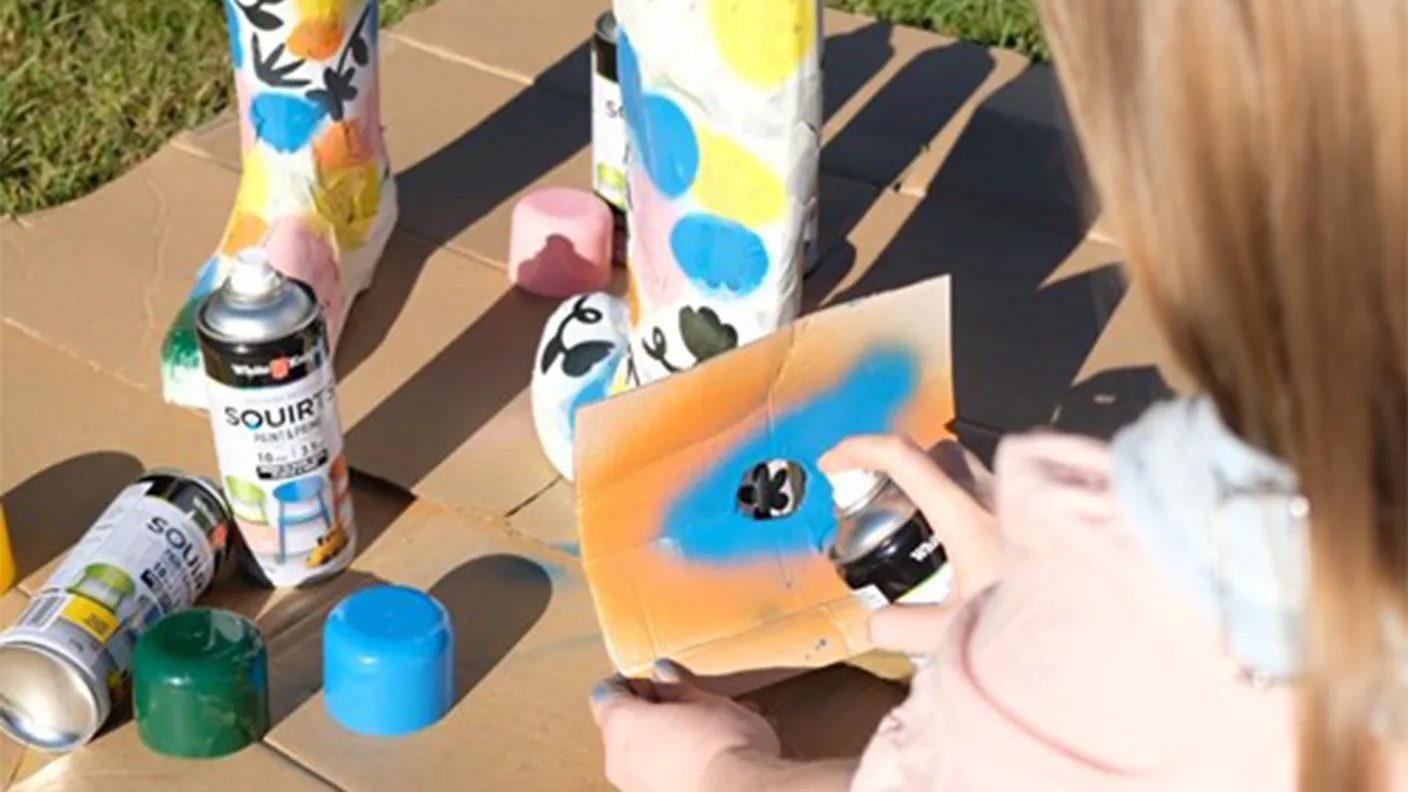 Person spray paints colorful boots using a stencil on cardboard, surrounded by cans and caps in outdoor setting.