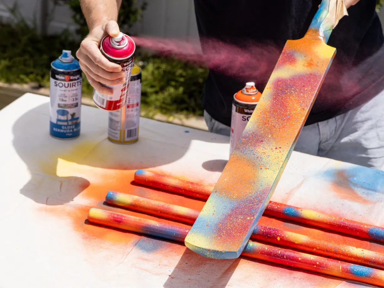 Person spray-painting a cricket bat with colorful splashes on a table covered in painted sticks and spray cans.