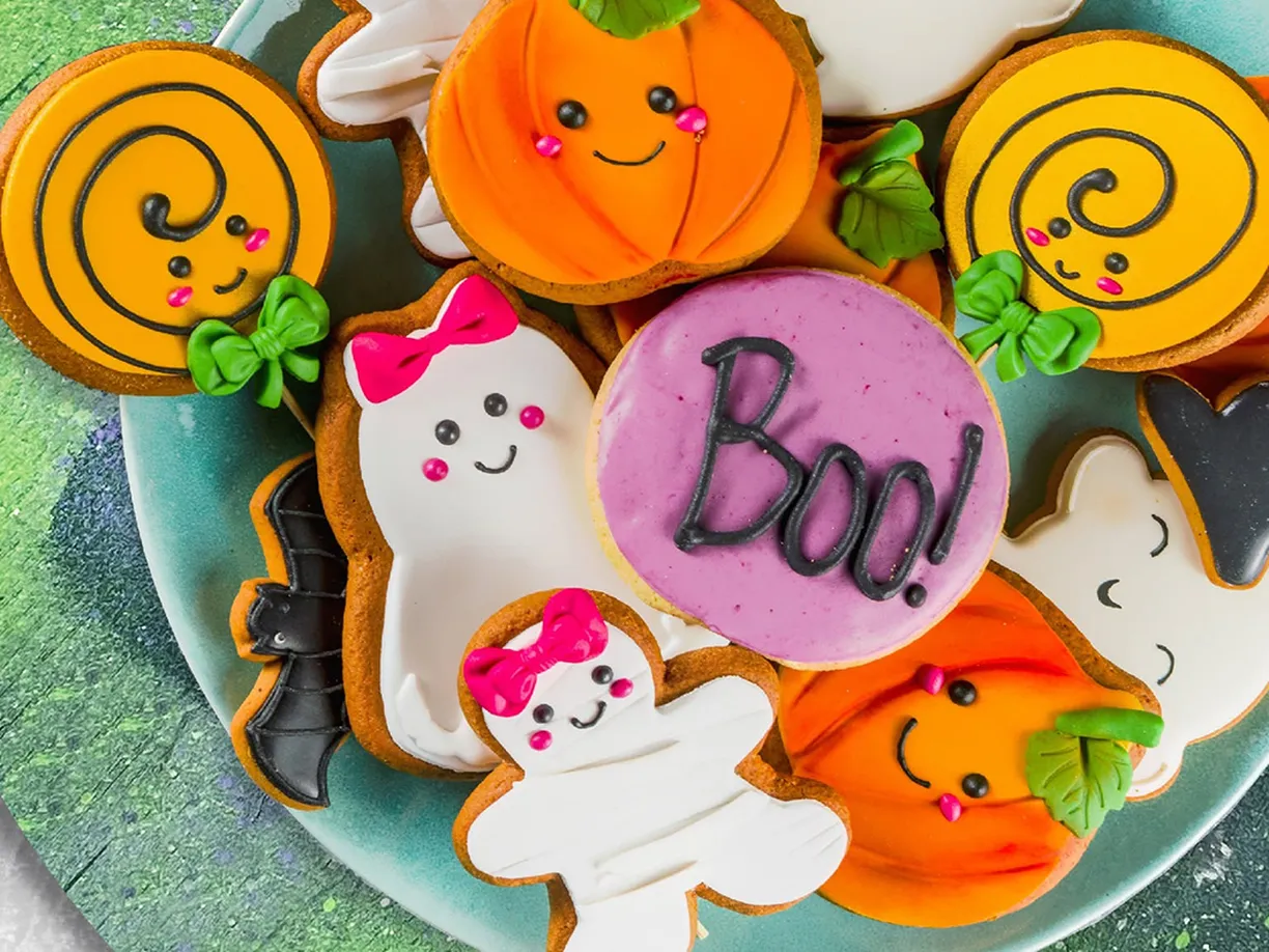A plate of Halloween-themed cookies, including pumpkins, ghosts, a cat, and a pink cookie with "Boo!" written on it.