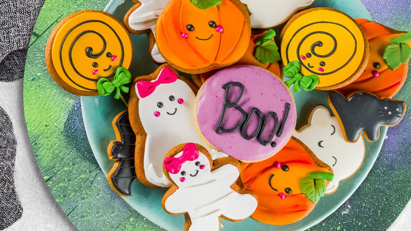 A plate of Halloween-themed cookies, including pumpkins, ghosts, a cat, and a pink cookie with "Boo!" written on it.