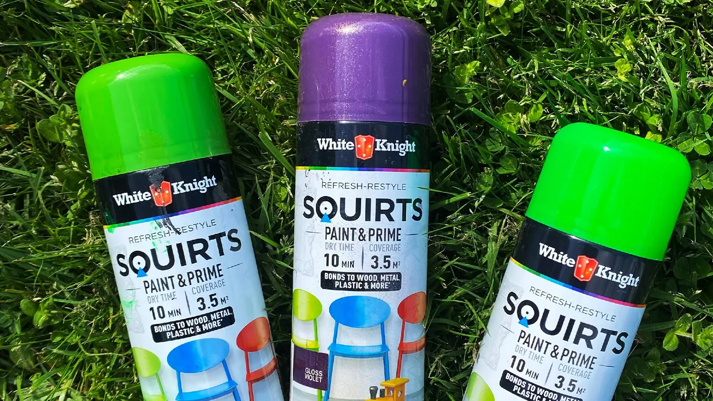 Three spray paint cans in green and purple lie on grass. Labels read "White Knight Squirts Paint & Prime," suitable for various surfaces.
