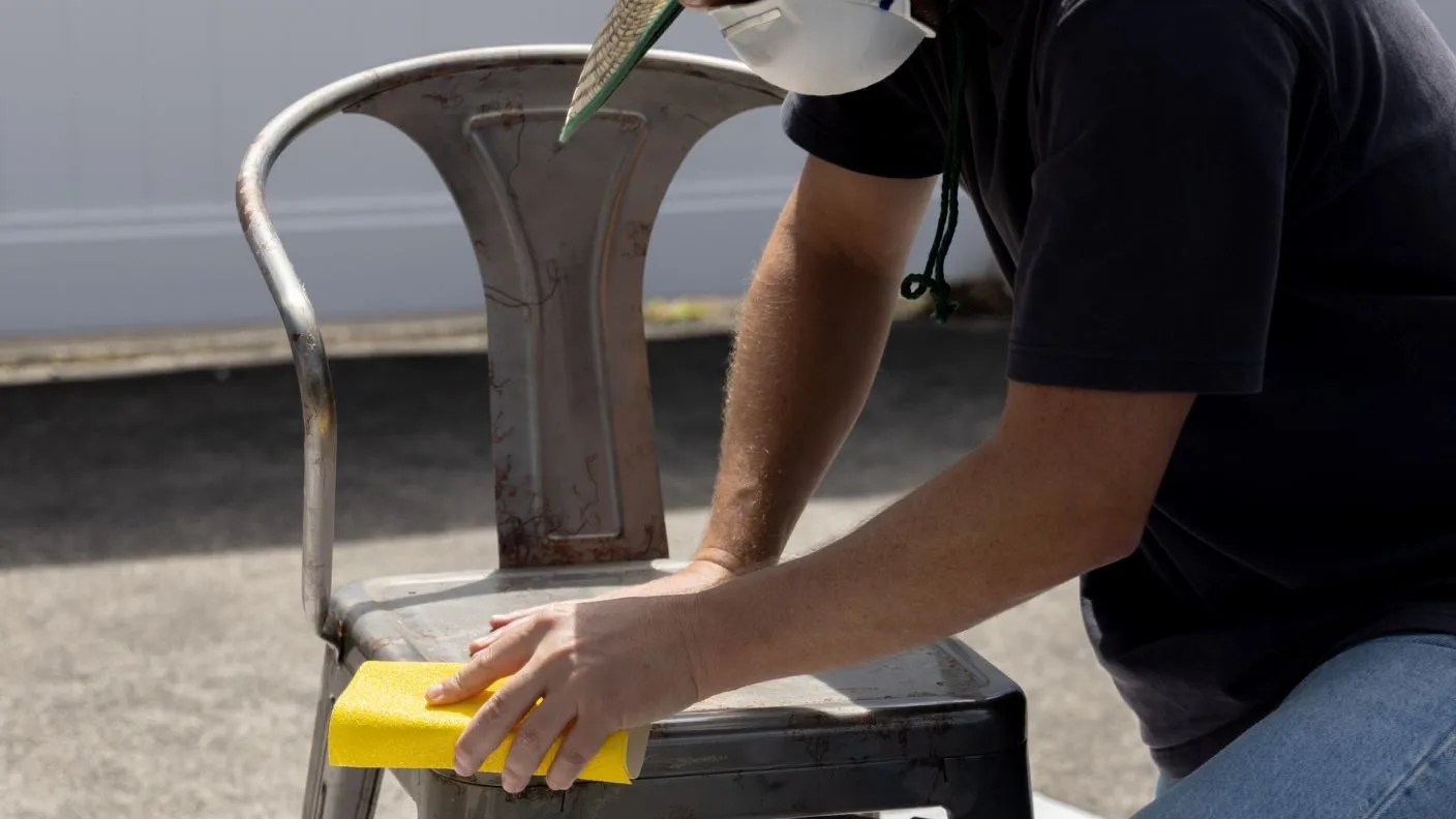 Man in a dust mask sanding a rusty metal outdoor chair with sandpaper to prep for painting