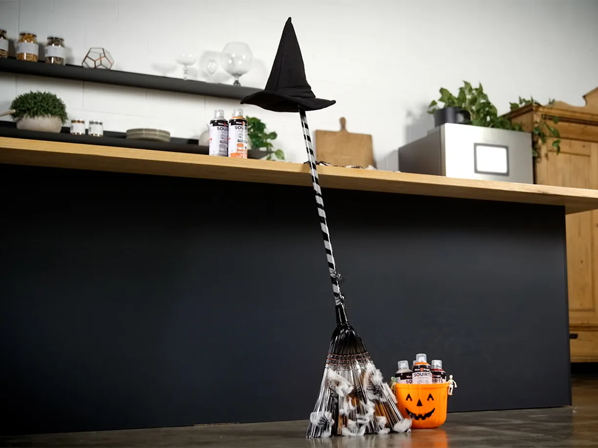A Halloween-themed broom with a witch's hat and black-and-white striped handle leans against a counter near a pumpkin bucket.