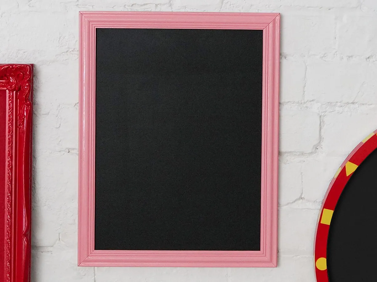 A pink-framed blackboard hangs on a white brick wall, flanked by a red frame on the left and a red-and-yellow patterned frame on the right.
