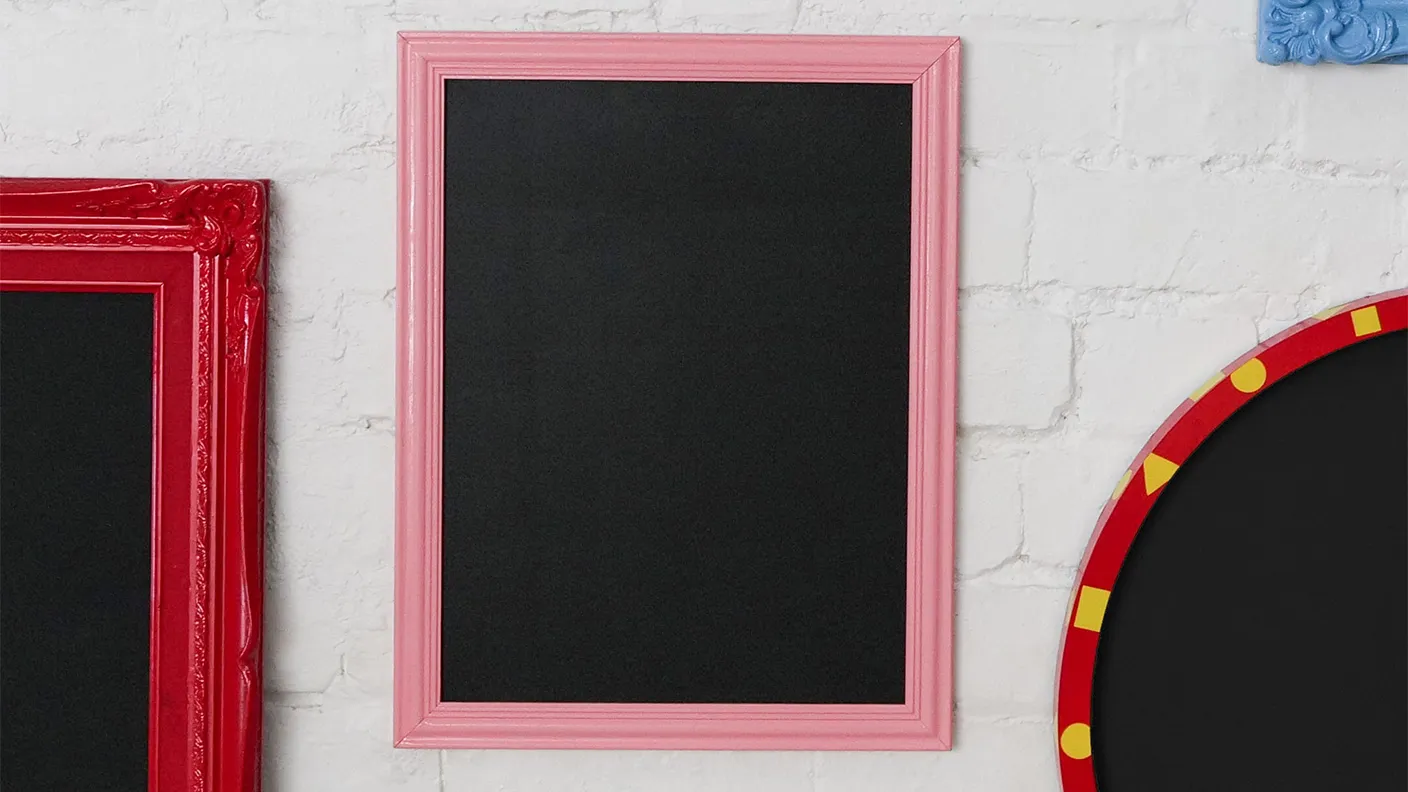 A pink-framed blackboard hangs on a white brick wall, flanked by a red frame on the left and a red-and-yellow patterned frame on the right.