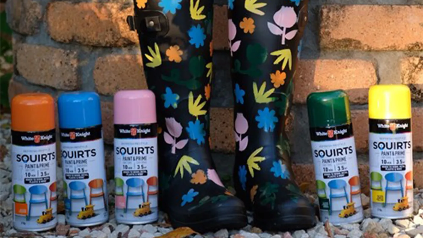 Patterned rain boots beside six colorful spray paint cans labeled "Squirts" on a stone and pebble background.