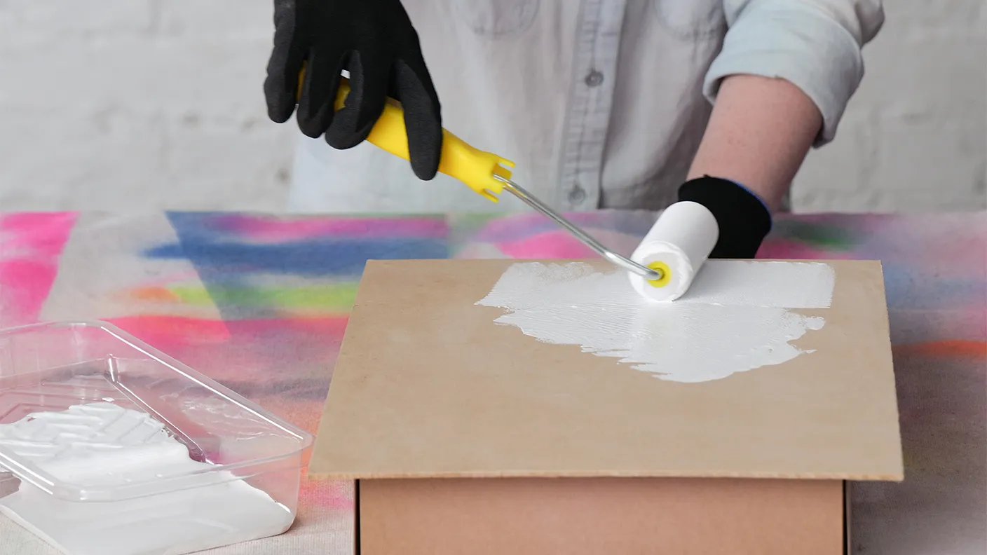 A person in black gloves uses a paint roller with white paint on a wooden surface, with a colorful background.