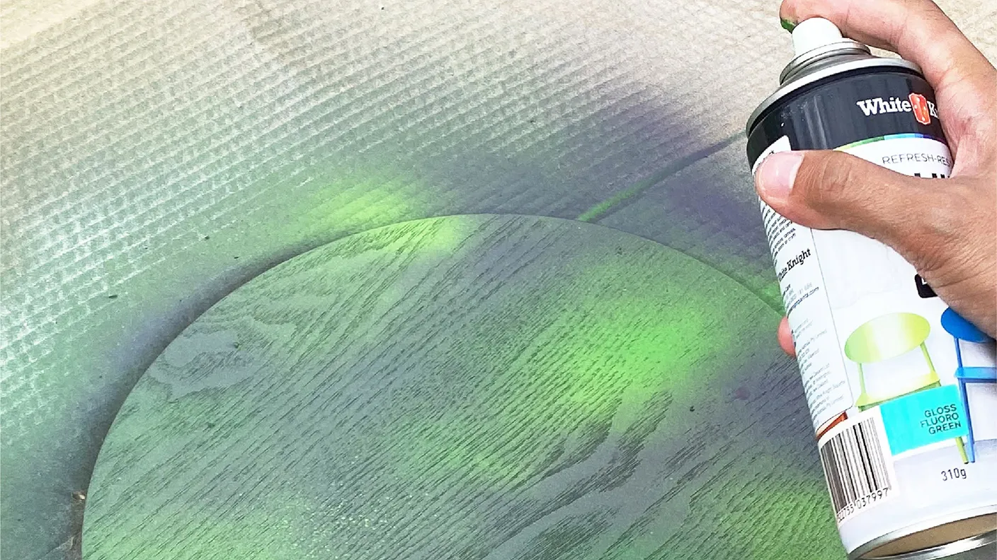 Hand holding a can of green spray paint, coloring a wooden surface with a round shape on a textured backdrop.