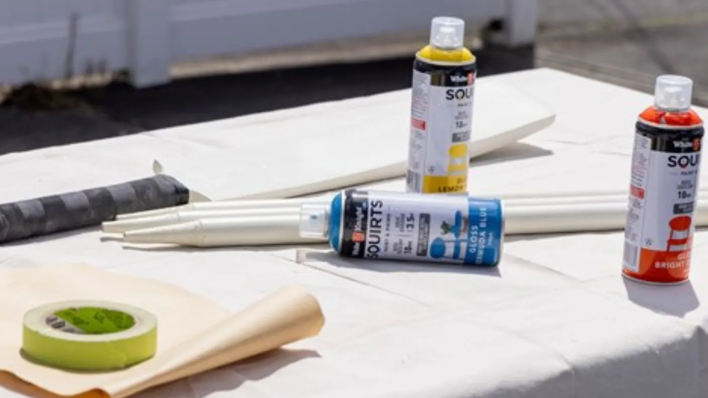 Spray paint cans in various colors on a table with masking tape, rolled paper, and a taped black rod, suggesting a painting project setup.