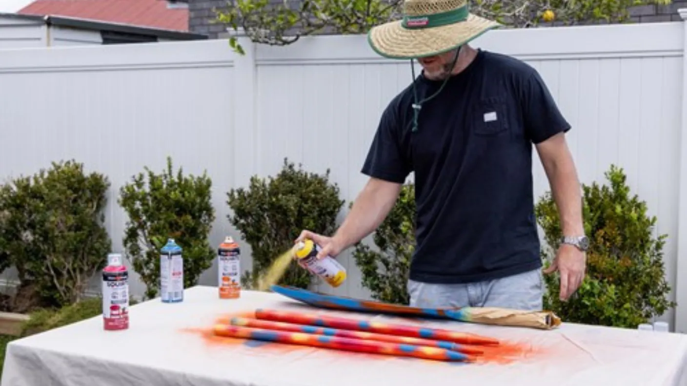 Man in a straw hat spray paints colorful designs on long objects placed on a table in a garden, surrounded by spray cans.