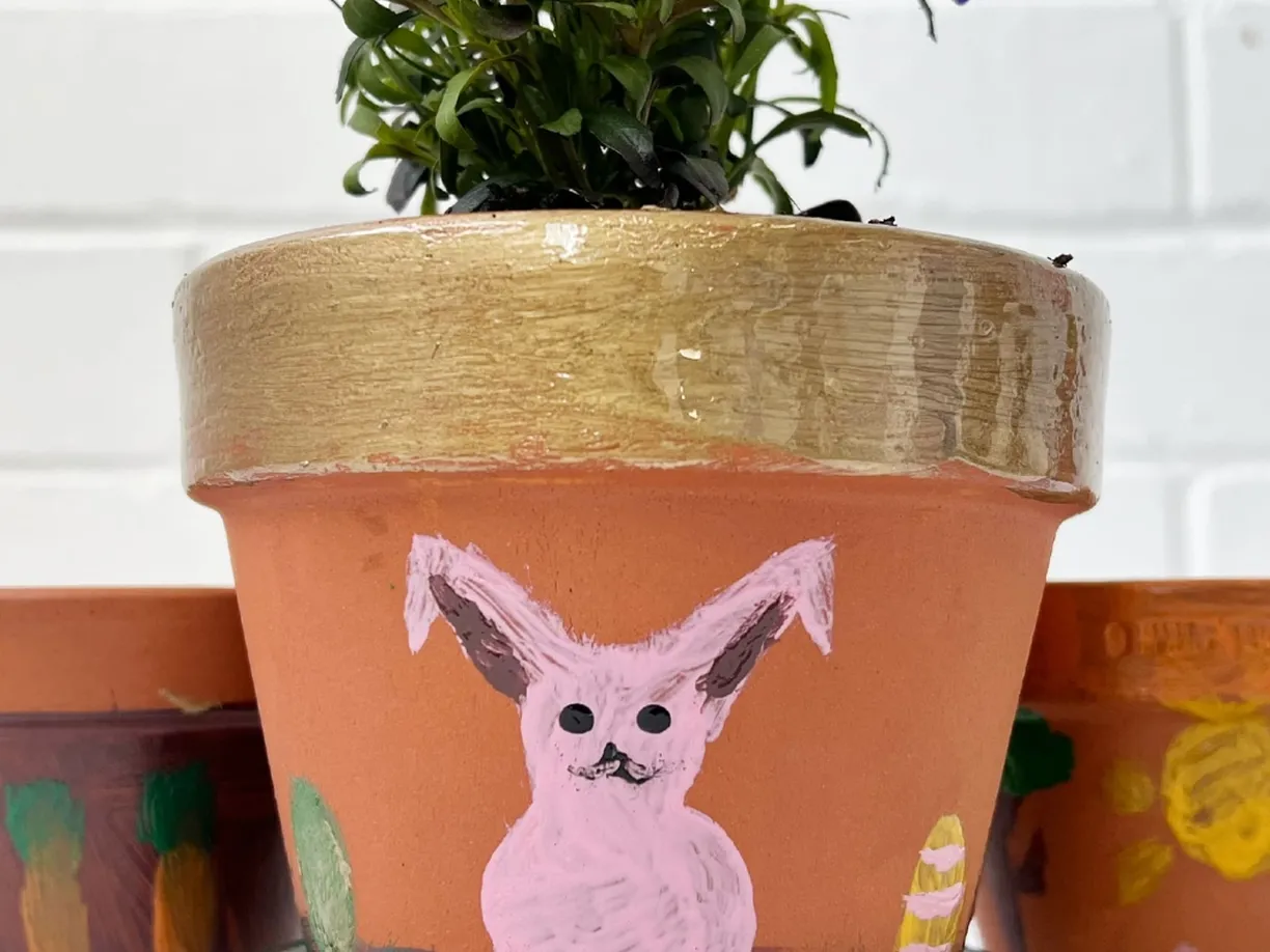 Clay pot with a gold-rimmed top and a painted pink rabbit, holding a plant, against a white brick wall background.