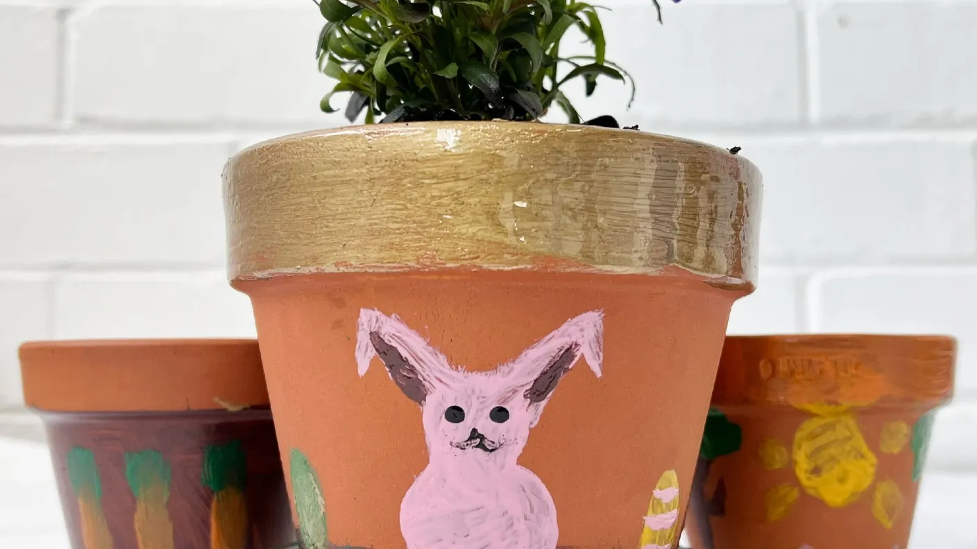 Clay pot with a gold-rimmed top and a painted pink rabbit, holding a plant, against a white brick wall background.