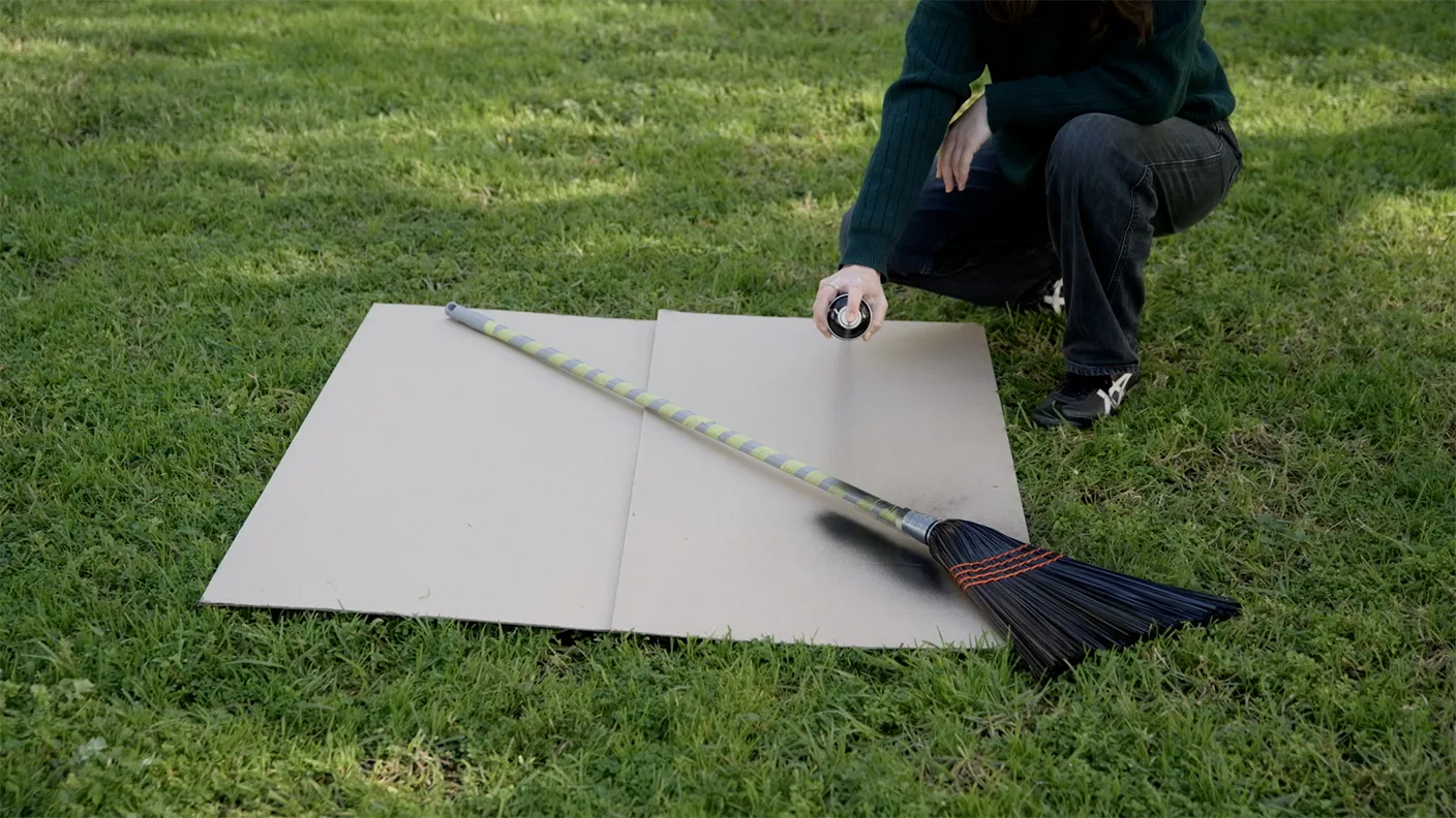 Person spray-painting a broom on a cardboard sheet placed on grass.