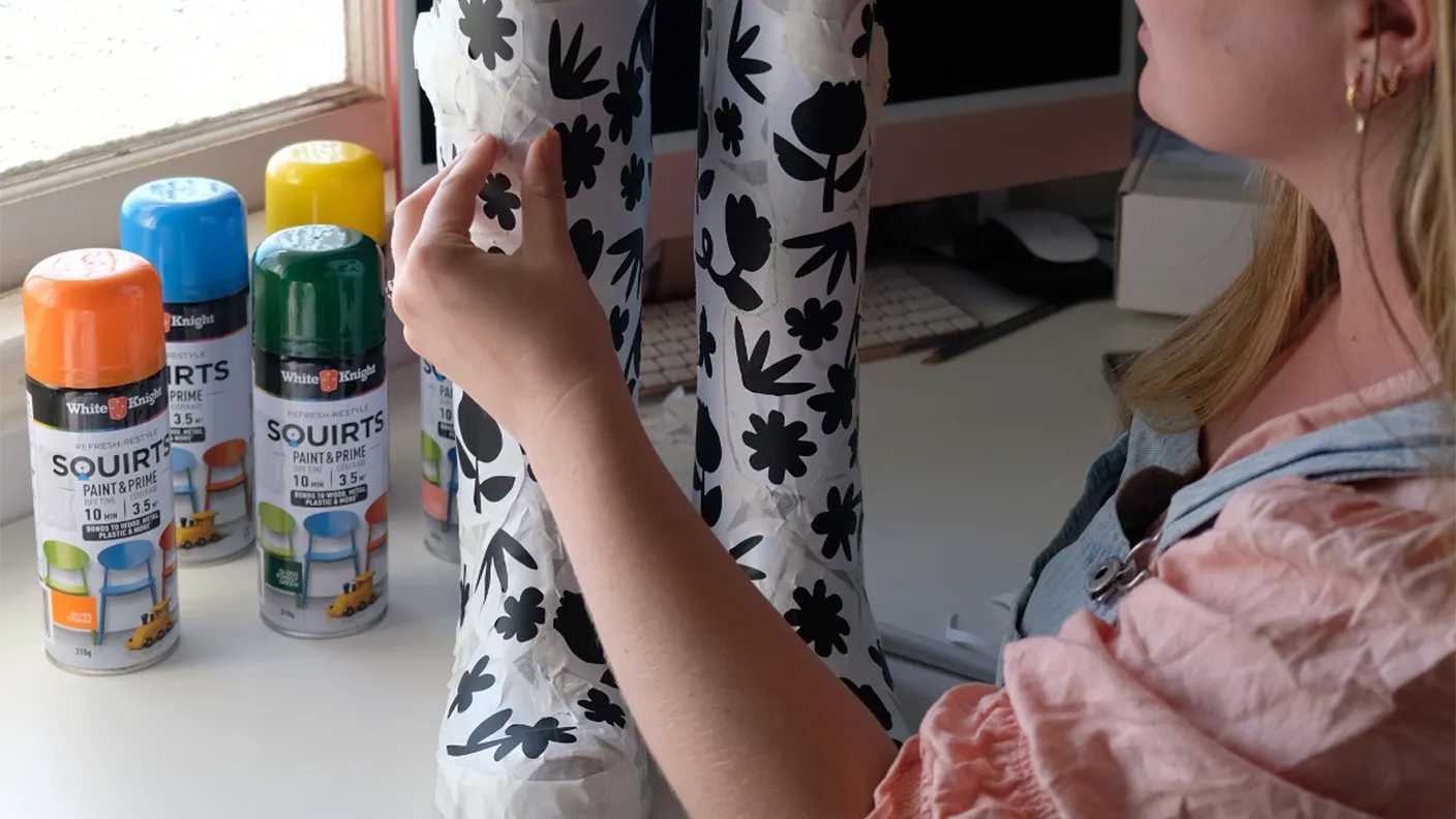 A person applies floral stencils to boots, with colorful spray paint cans nearby on a desk in a craft room.