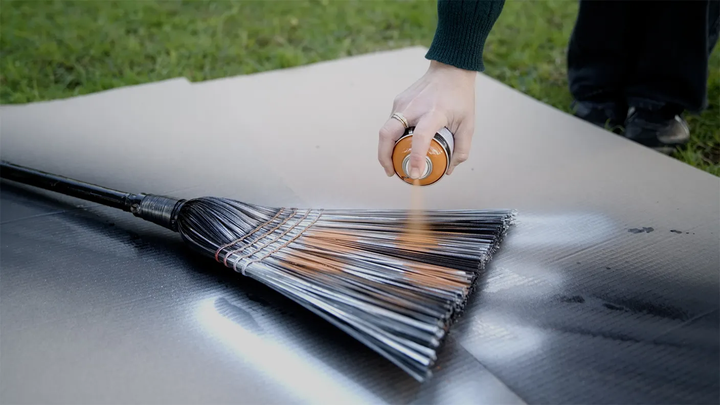 Hand spray-painting a broom with orange paint on cardboard outdoors.