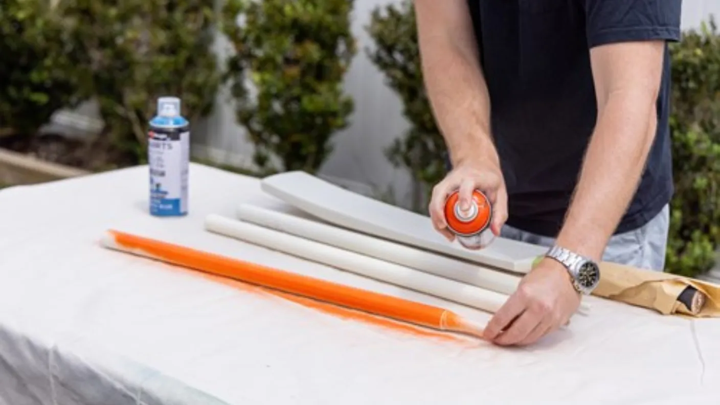 Person spray-painting a white object orange on a table outdoors, with other items and a can of spray paint nearby.