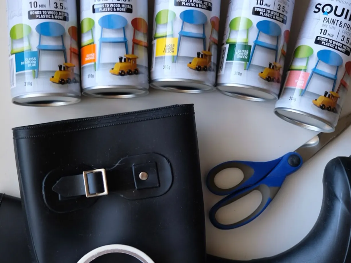 Black rubber boots, blue-handled scissors, and five cans of colorful "Squirts" paint on a white surface.