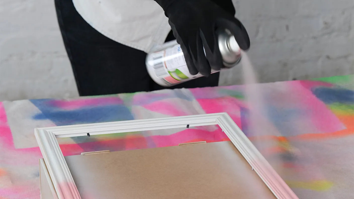 Person spray-painting a white picture frame over a colorful, abstract background while wearing black gloves.