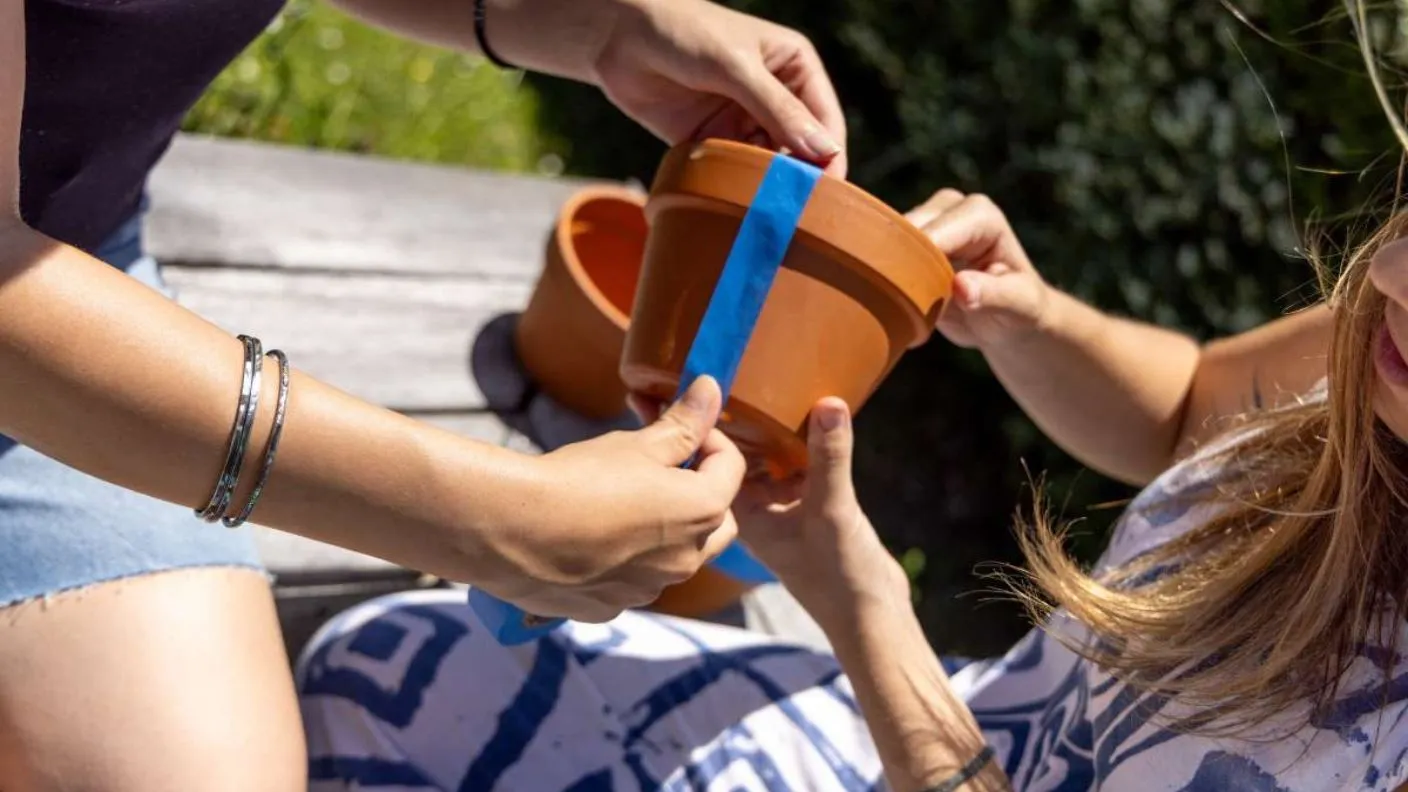 A person tapes up a terracotta pot on a drop sheet, with a gold spray-painted pot and a plain terracotta pot nearby ready for customising with White Knight Squirts