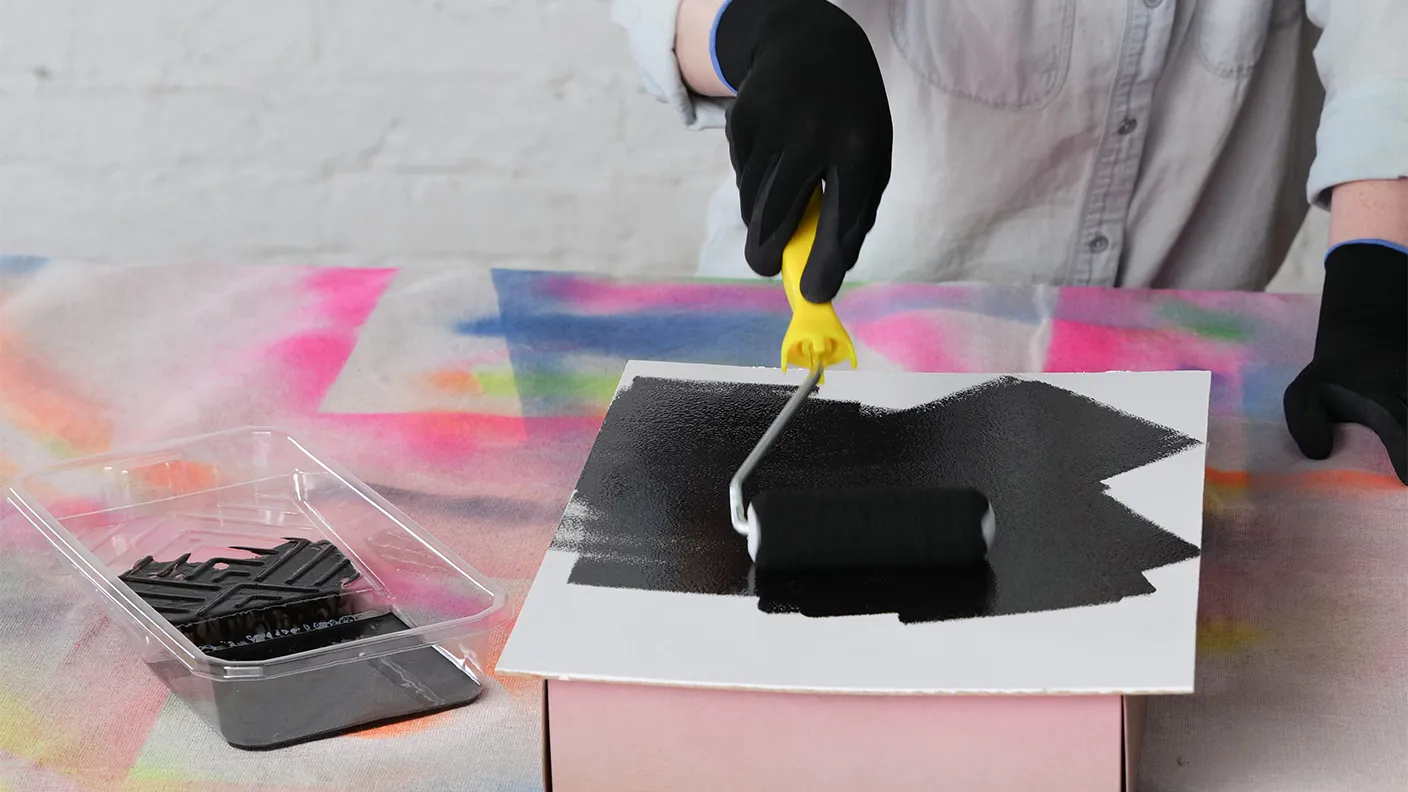 Person wearing black gloves uses a paint roller to apply black paint on white paper over a colorful tablecloth.