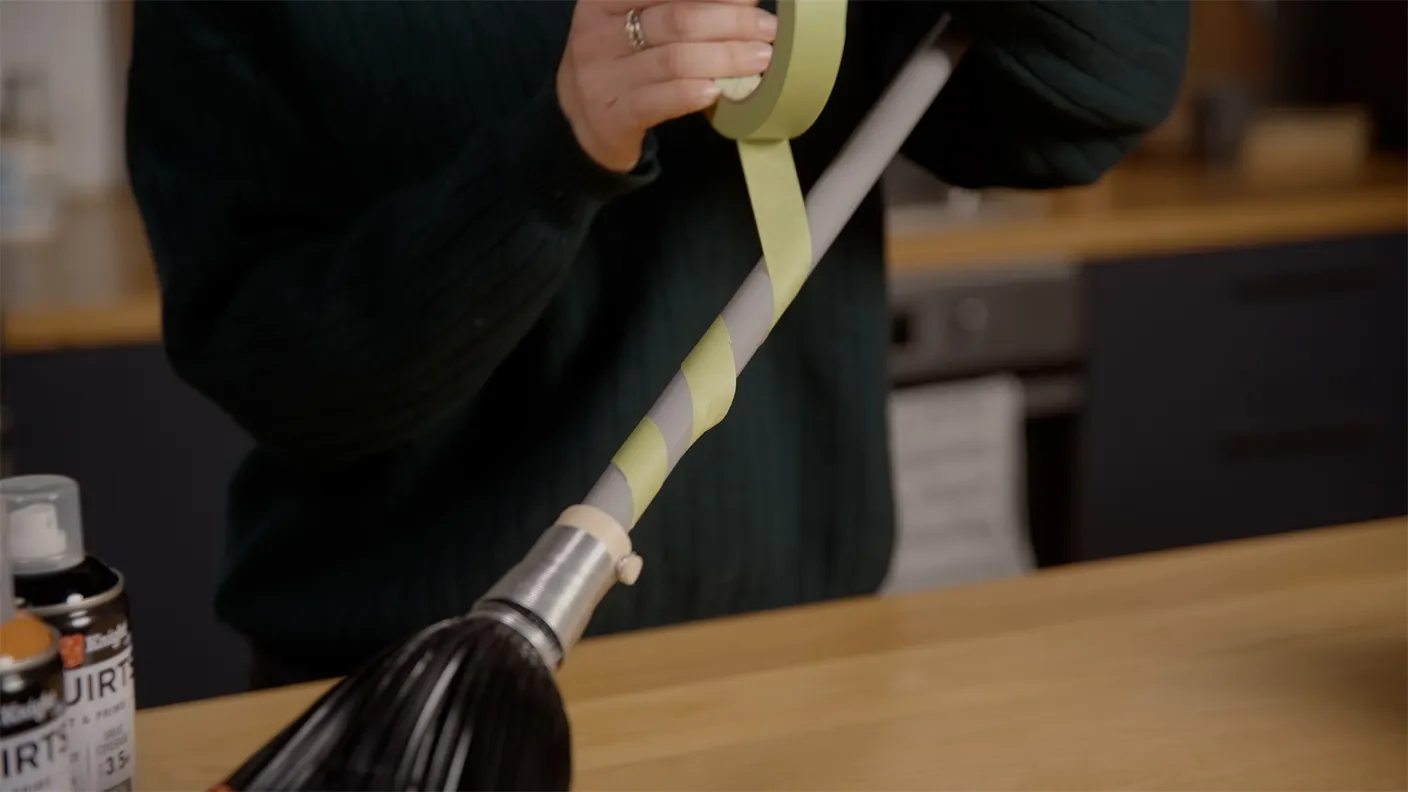 Person wrapping yellow tape around a broom handle, with spray cans visible on the table in a kitchen setting.