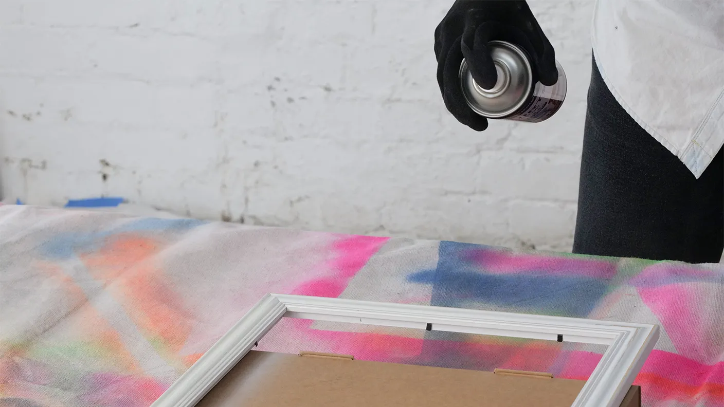 Person in gloves spray painting a white picture frame on colorful fabric against a white brick wall.