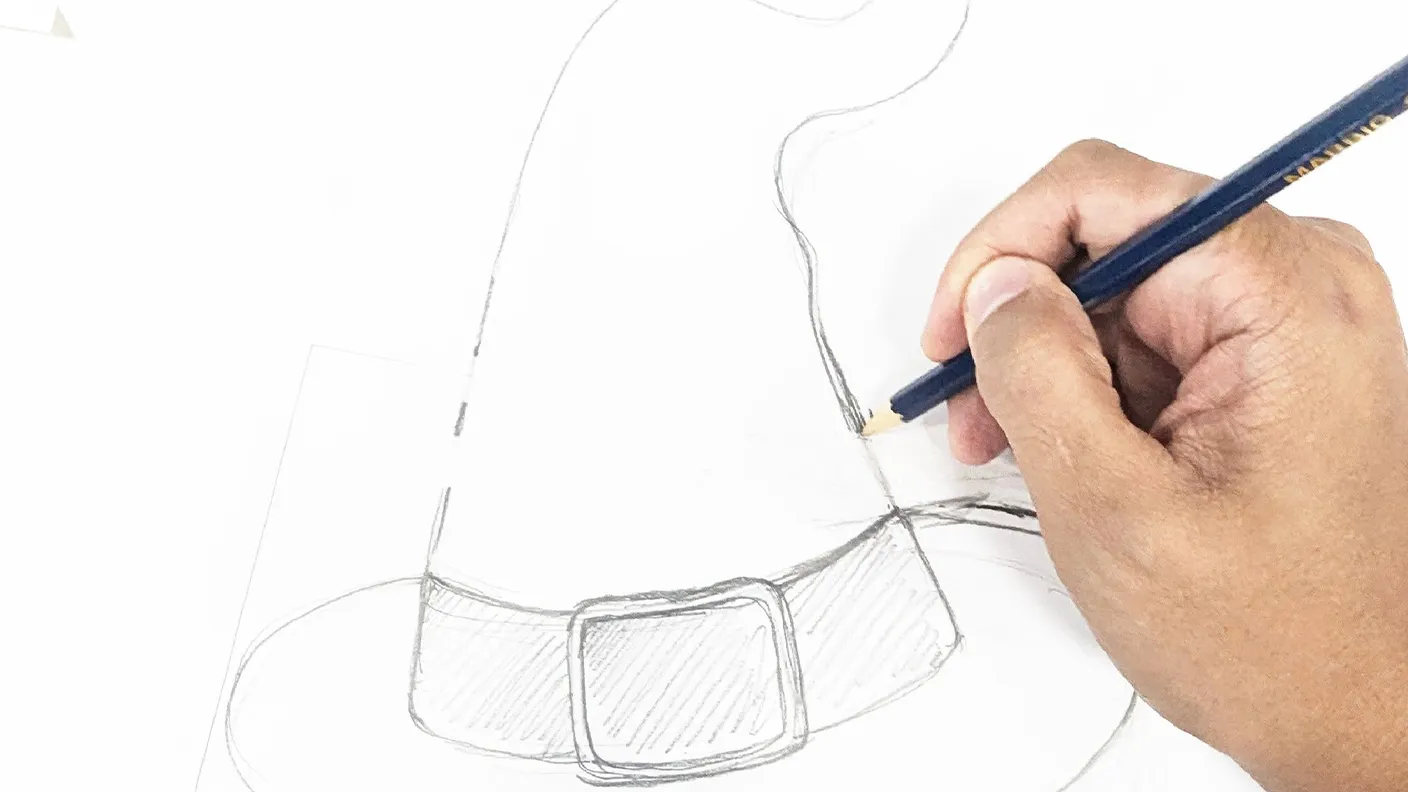 Hand sketching a witch hat with a pencil on white paper, focusing on the belt detail.