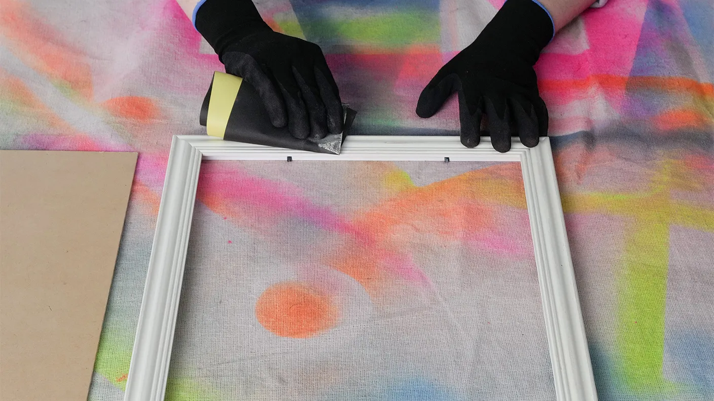 Hands wearing black gloves sanding a white picture frame on a colorful background.