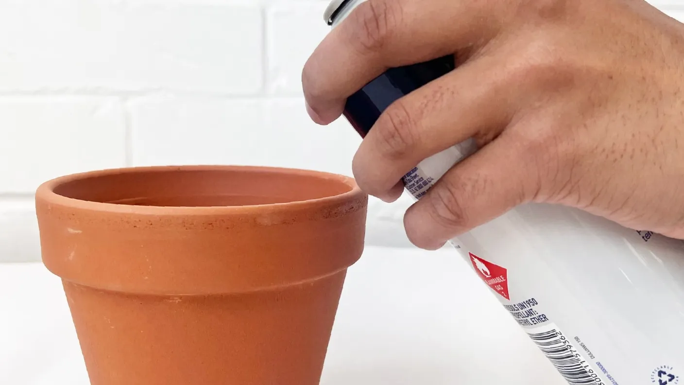 Hand using a spray can to paint a terracotta pot against a white background.